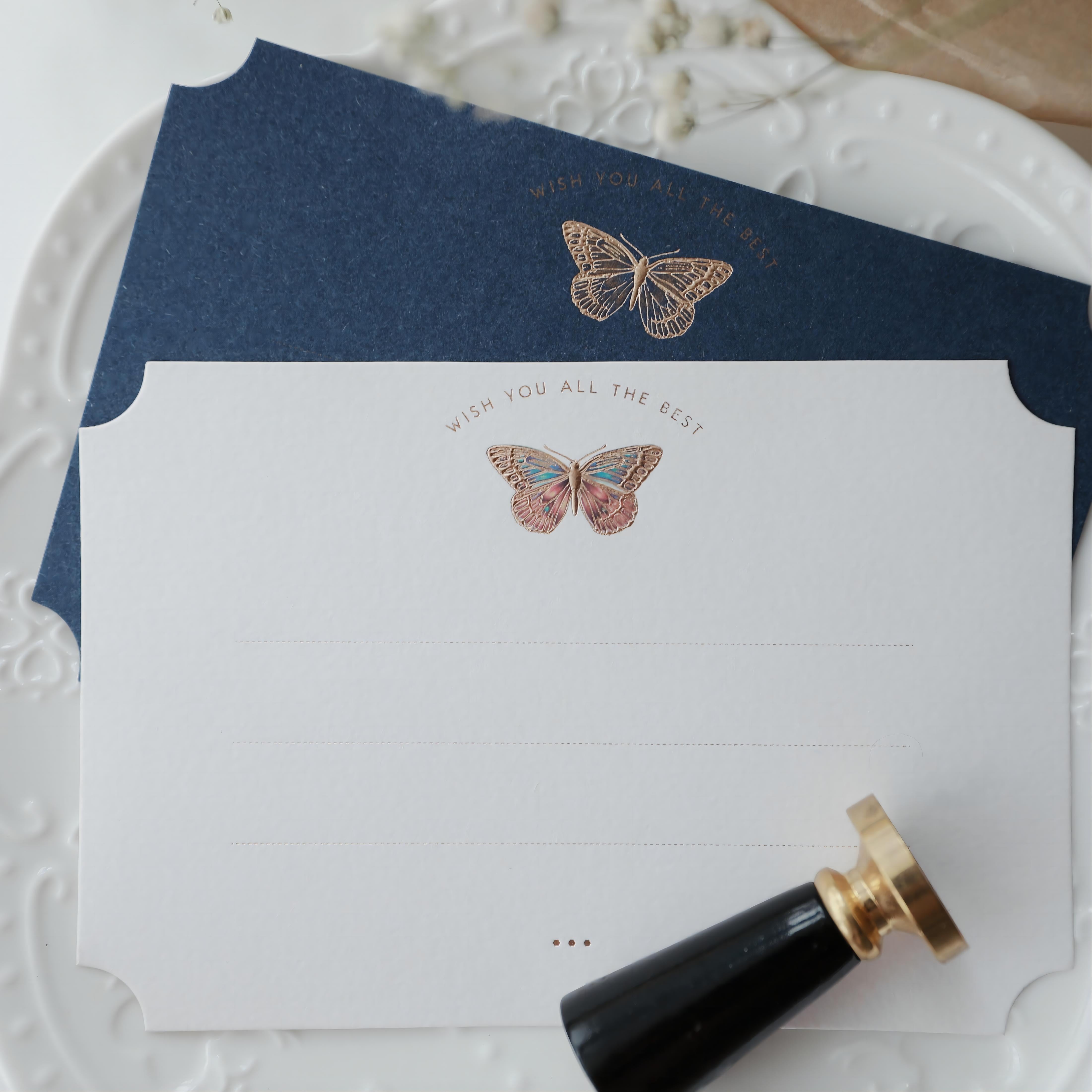 "Dreamy Blessings" greeting card set featuring two elegant cards. The first card is premium white with an embossed butterfly design in gold foil that reads "Wish You All The Best," and the second card is a deep navy blue with a similar gold foil butterfly. Both cards rest elegantly on a decorative white plate with embossed patterns, accompanied by a classic black and gold wax seal stamp. This set is perfect for conveying warm wishes on special occasions. Made from eco-friendly materials.