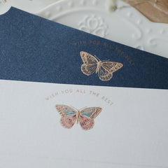 "Dreamy Blessings" greeting card set featuring two elegant cards, one in premium white and the other in deep navy blue, each with a gold foil embossed butterfly and the message "Wish You All The Best." The cards are displayed on a decorative white plate with embossed patterns, highlighting their refined design. Perfect for expressing heartfelt wishes on special occasions. Made from eco-friendly materials.