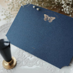 "Dreamy Blessings" greeting card in deep navy blue, featuring a gold foil embossed butterfly with the message "Wish You All The Best." The card is elegantly placed on a decorative white plate with intricate embossed patterns and surrounded by delicate baby's breath flowers. A black and gold wax seal stamp is positioned nearby, enhancing the card's sophisticated and elegant appearance. This card is ideal for conveying heartfelt wishes on special occasions. Made from eco-friendly materials.