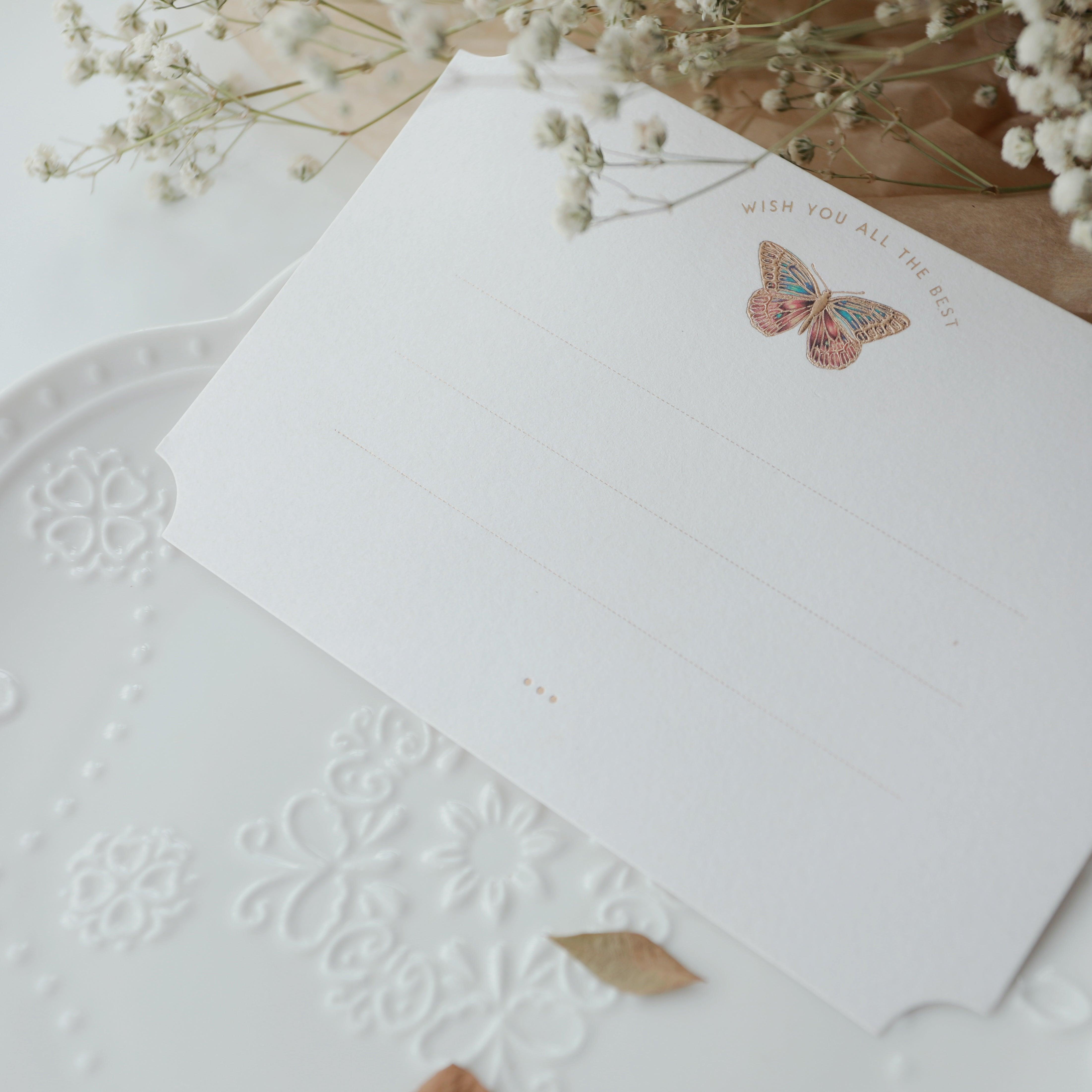 "Dreamy Blessings" greeting card in premium white, featuring a delicate gold foil embossed butterfly with the message "Wish You All The Best." The card is beautifully placed on an embossed white plate adorned with intricate floral patterns, with a few dried leaves adding a touch of nature. Surrounded by delicate baby's breath flowers, this card is perfect for expressing sincere wishes on special occasions. Made from eco-friendly materials.