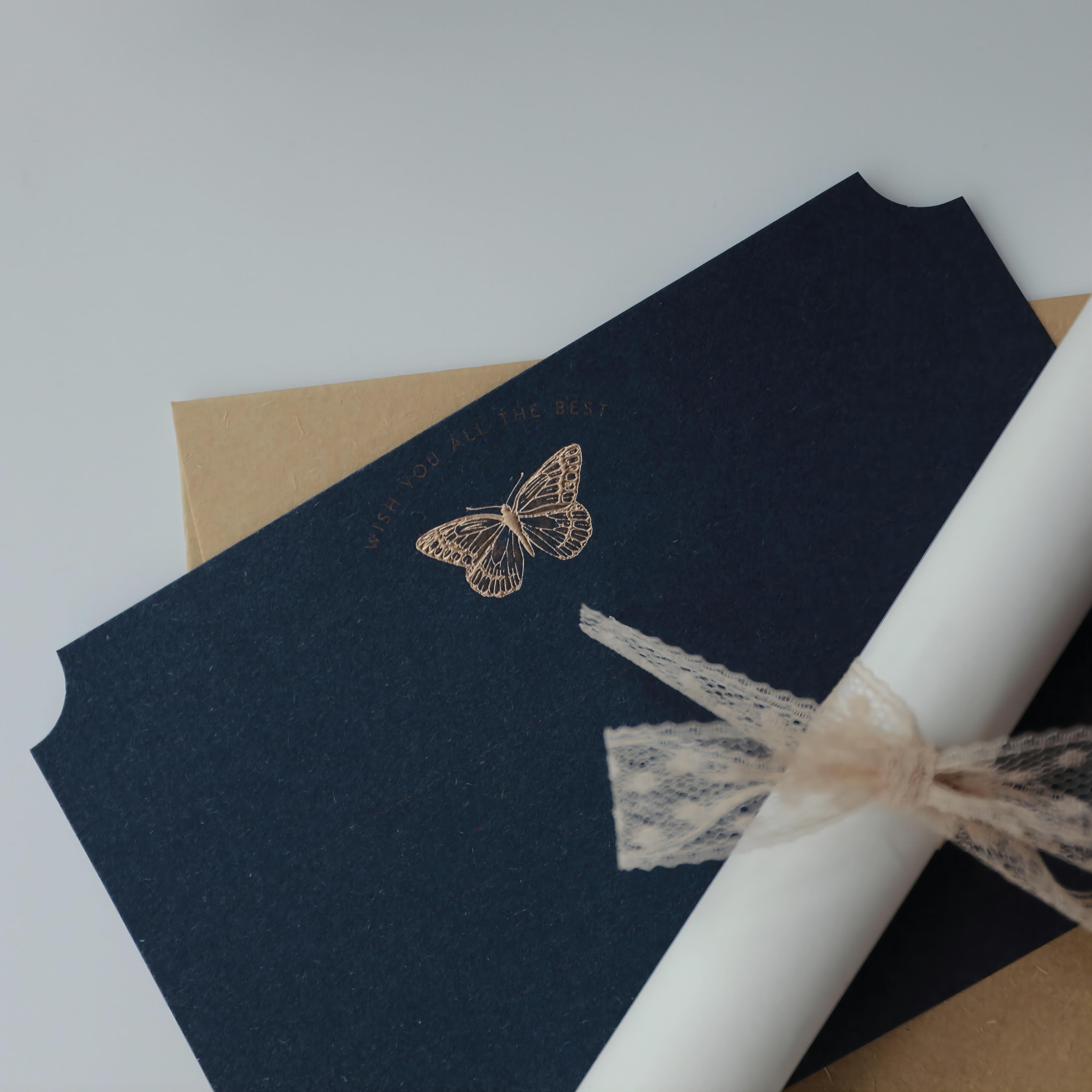 "Dreamy Blessings" greeting card in deep navy blue, featuring a gold foil embossed butterfly with the message "Wish You All The Best." The card is accompanied by a natural kraft envelope and a rolled-up parchment tied with a delicate lace ribbon, creating a charming and elegant presentation. This card set is perfect for conveying heartfelt wishes on special occasions. Made from eco-friendly materials.