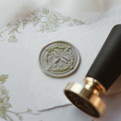 "Firefly Whisper" wax seal stamp featuring an intricately designed seal with a delicate firefly motif. The seal is elegantly pressed onto a textured white paper, surrounded by floral-patterned fabric. The accompanying black handle and gold head stamp lie nearby, highlighting the craftsmanship and elegance of this unique seal. Perfect for adding a touch of sophistication to letters or invitations. Made with high-quality materials.