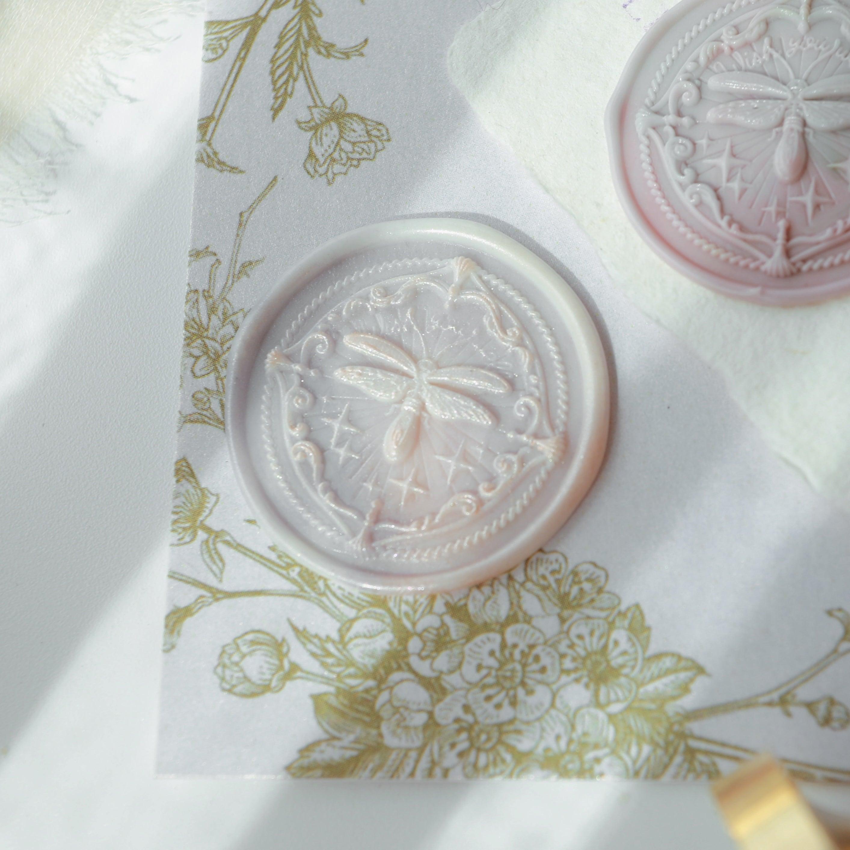 "Firefly Whisper" wax seals featuring an intricately designed seal with a delicate firefly motif. The seals are elegantly displayed on textured paper with floral patterns, capturing the essence of nature and craftsmanship. The soft, translucent pink color of the wax adds a touch of elegance and sophistication. Perfect for enhancing the charm of letters or invitations. Crafted from high-quality materials.