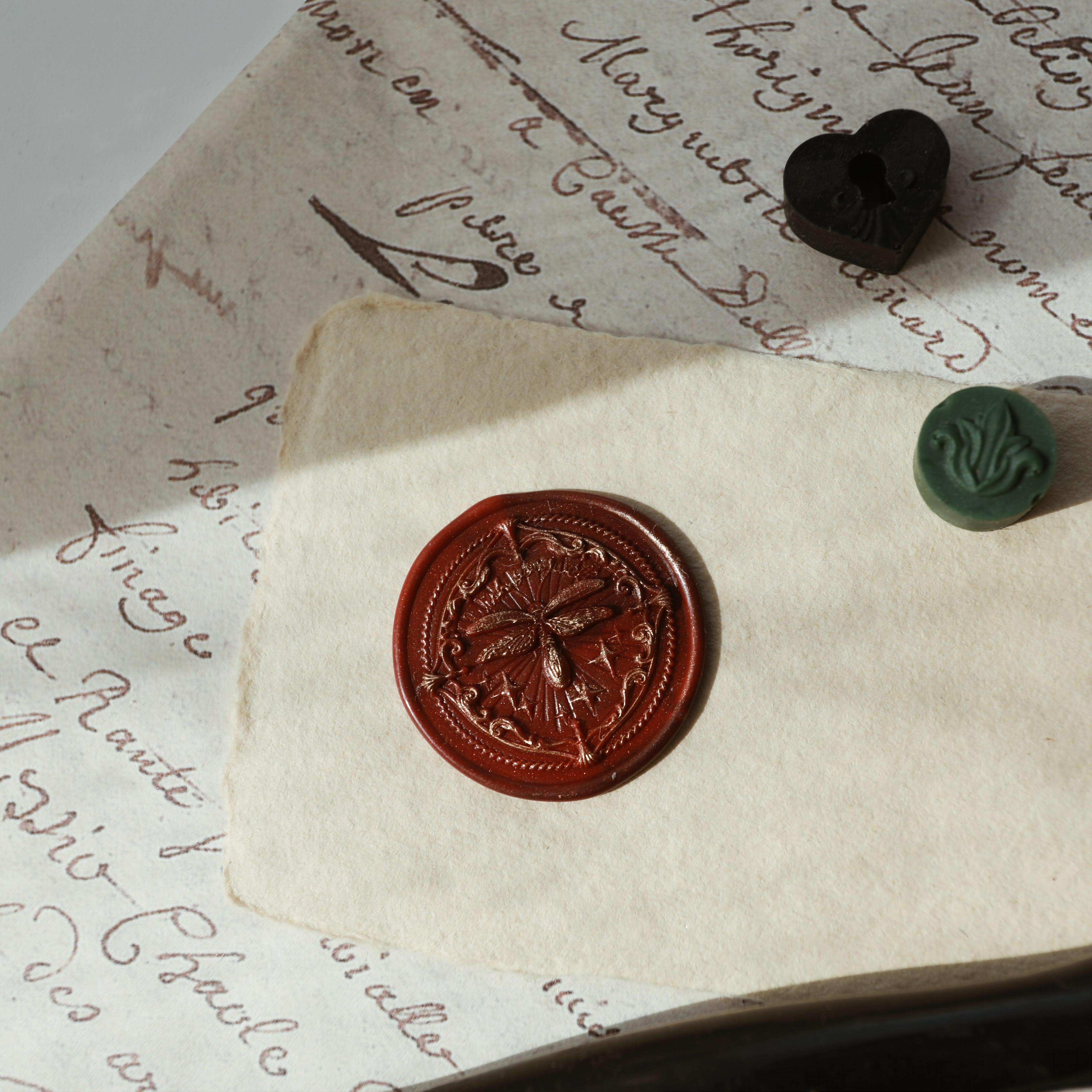 "Firefly Whisper" wax seal featuring an intricately designed seal with a delicate firefly motif. The seal is embossed in a rich brown wax, creating a classic and vintage look. It is displayed on a piece of aged parchment, surrounded by calligraphic handwriting, along with a black heart-shaped wax bead and a green round wax bead. This setup evokes a sense of timeless elegance and sophistication, perfect for adding a personal touch to letters or invitations.