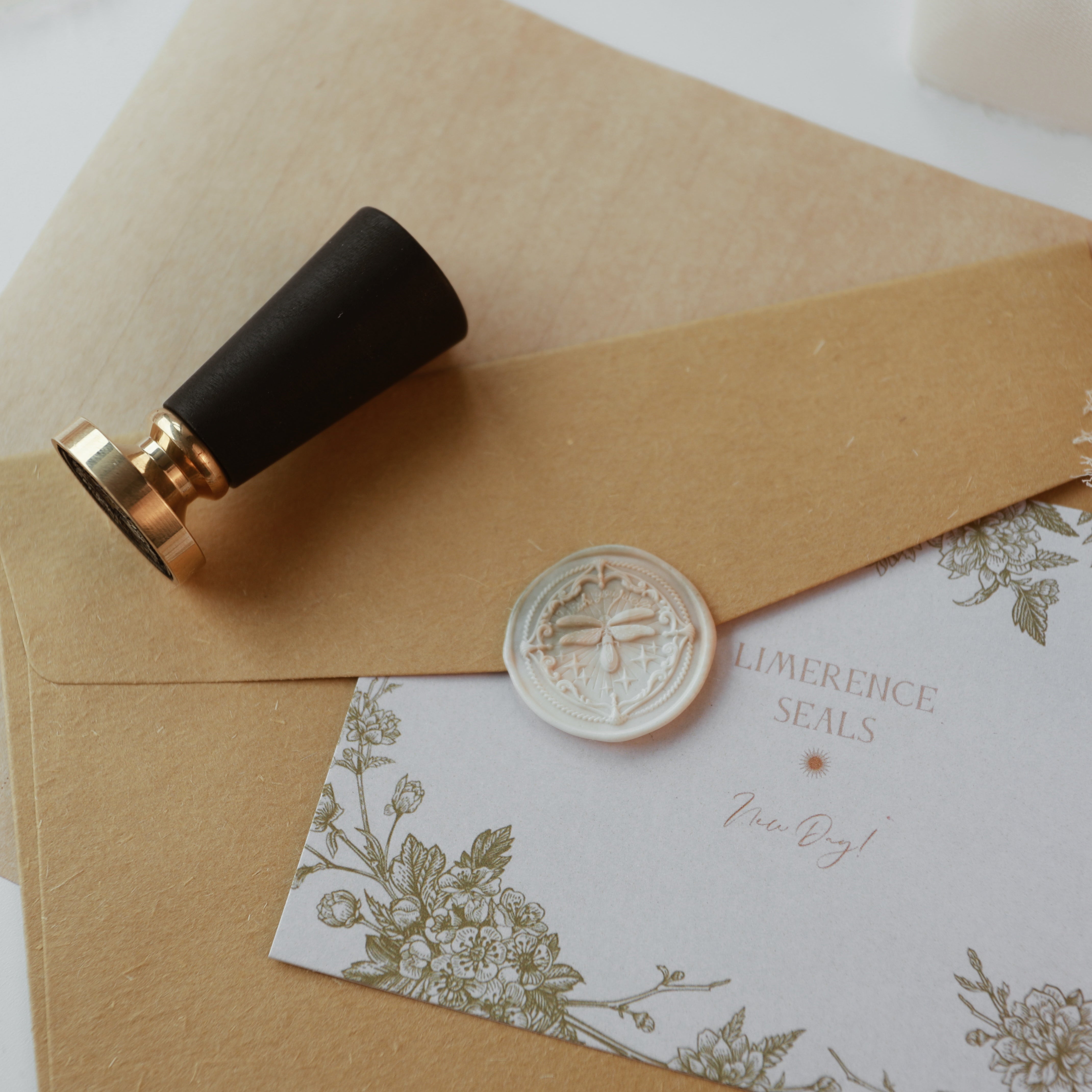 "Firefly Whisper" wax seal stamp, featuring an elegantly designed seal with a firefly motif. The seal is pressed onto a natural kraft envelope, accompanied by a decorative card with floral designs and the text "LIMERENCE SEALS." A black-handled, gold-headed stamp lies nearby, showcasing the seal's craftsmanship. This setup offers a rustic and sophisticated feel, ideal for personalizing invitations or correspondence. Crafted from high-quality materials.