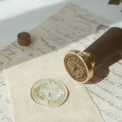 "Firefly Whisper" wax seal stamp featuring a detailed firefly motif. The seal is pressed into a marbled, multicolored wax, creating a striking impression on aged parchment. A black-handled, gold-headed stamp is positioned nearby, showcasing exquisite craftsmanship. Alongside a brown wax bead resting on vintage handwritten script, this setup exudes a timeless elegance perfect for personalizing letters or invitations.