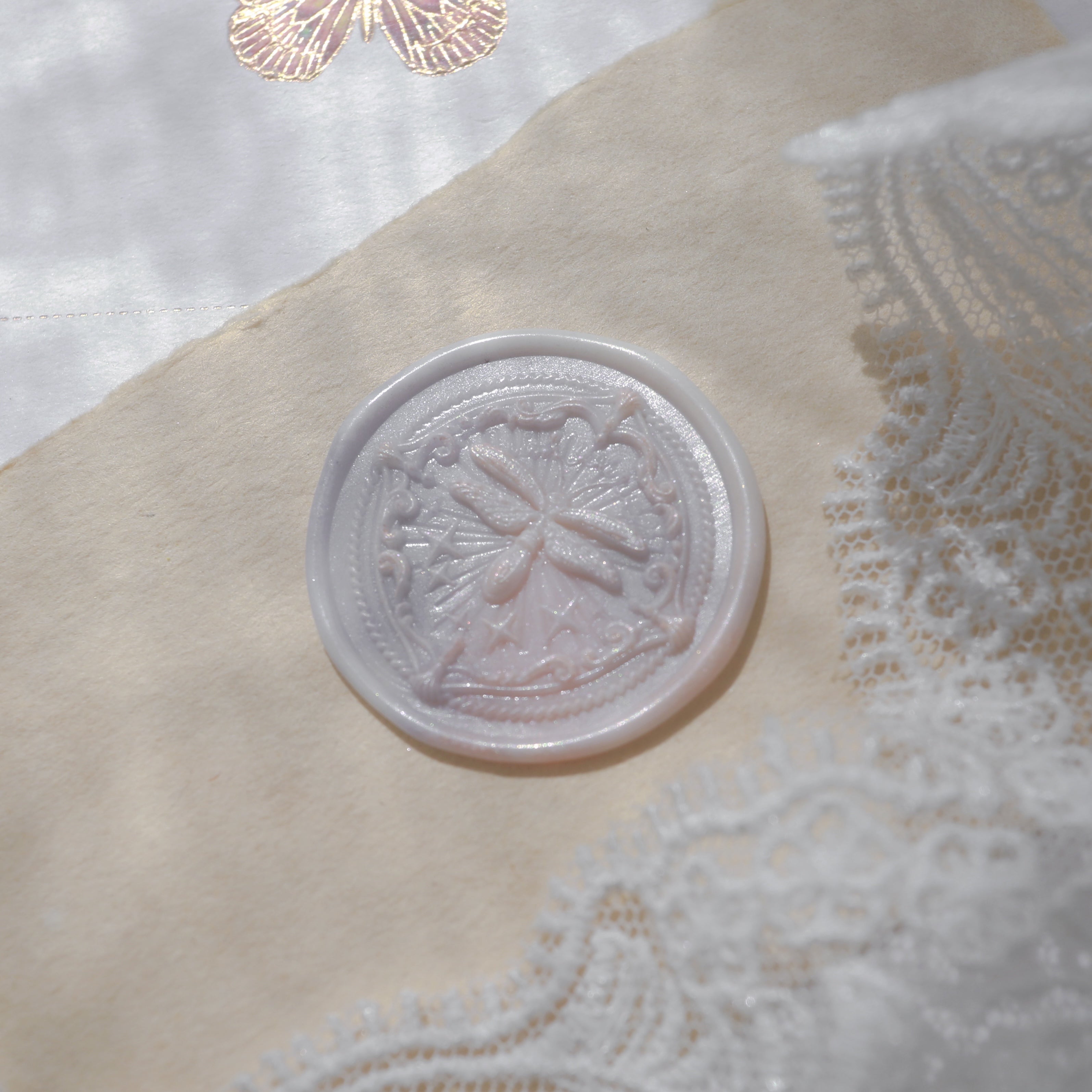 "Firefly Whisper" wax seal featuring an intricate firefly design. The seal is beautifully pressed into a marbled wax, creating an elegant impression on aged parchment. It is placed next to delicate lace fabric and a card with a gold foil butterfly motif, enhancing the scene's sophistication and charm, perfect for adding a personal touch to letters or invitations.