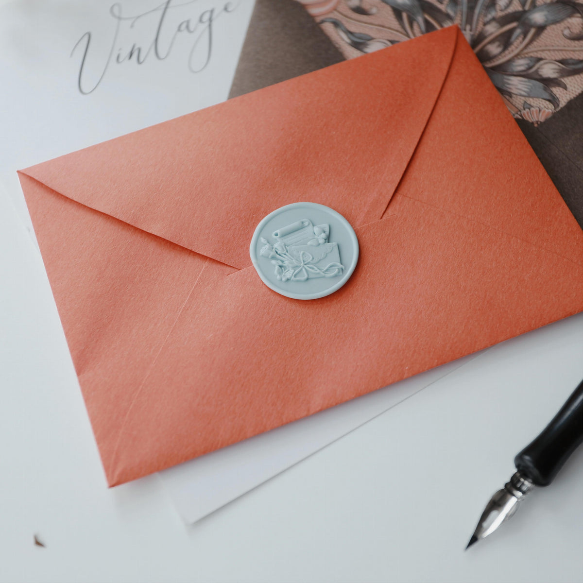 An elegant coral-orange envelope from the "Floral Elegance" series by Limerence Seals, sealed with a detailed light blue wax seal. The seal features an intricate design of a fountain pen, an envelope, and flowers tied with a ribbon. The envelope is set against a background of vintage-style artwork and a calligraphy sheet labeled "Vintage." A classic black fountain pen with a silver nib lies nearby, enhancing the timeless and artistic theme of the scene.