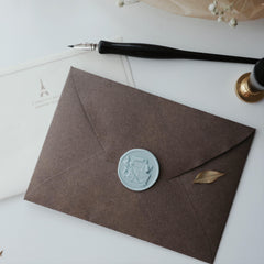 A sophisticated Dawn Brown envelope from the "Floral Elegance" series by Limerence Seals, featuring a beautifully detailed light blue wax seal. The wax seal showcases an intricate design of a fountain pen, an envelope, and flowers tied with a ribbon. A black fountain pen with a silver nib and a golden wax seal stamp are nearby, complementing the elegant aesthetic. The background includes a delicate sheet of stationery with an Eiffel Tower motif and French script, adding a touch of Parisian charm to the comp