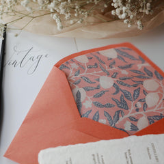 "Floral Elegance" vintage artistic envelope in a coral orange color, featuring a delicate floral and leaf pattern lining in soft pink and navy blue. The envelope is elegantly displayed on a surface with vintage calligraphy that reads "Vintage," alongside sprigs of baby's breath flowers and textured white paper, creating an artistic and nostalgic atmosphere. Ideal for adding a touch of elegance and sophistication to your letters. Made from eco-friendly materials.