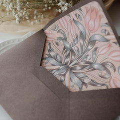 "Floral Elegance" vintage artistic envelope in a dawn brown color, featuring an intricate floral pattern with pink and blue-grey tones on the inside lining. The envelope is beautifully positioned against a soft background with delicate baby's breath flowers and a white plate with a subtle embossed edge. This design exudes a classic and elegant charm, perfect for special occasions. Made from eco-friendly materials.