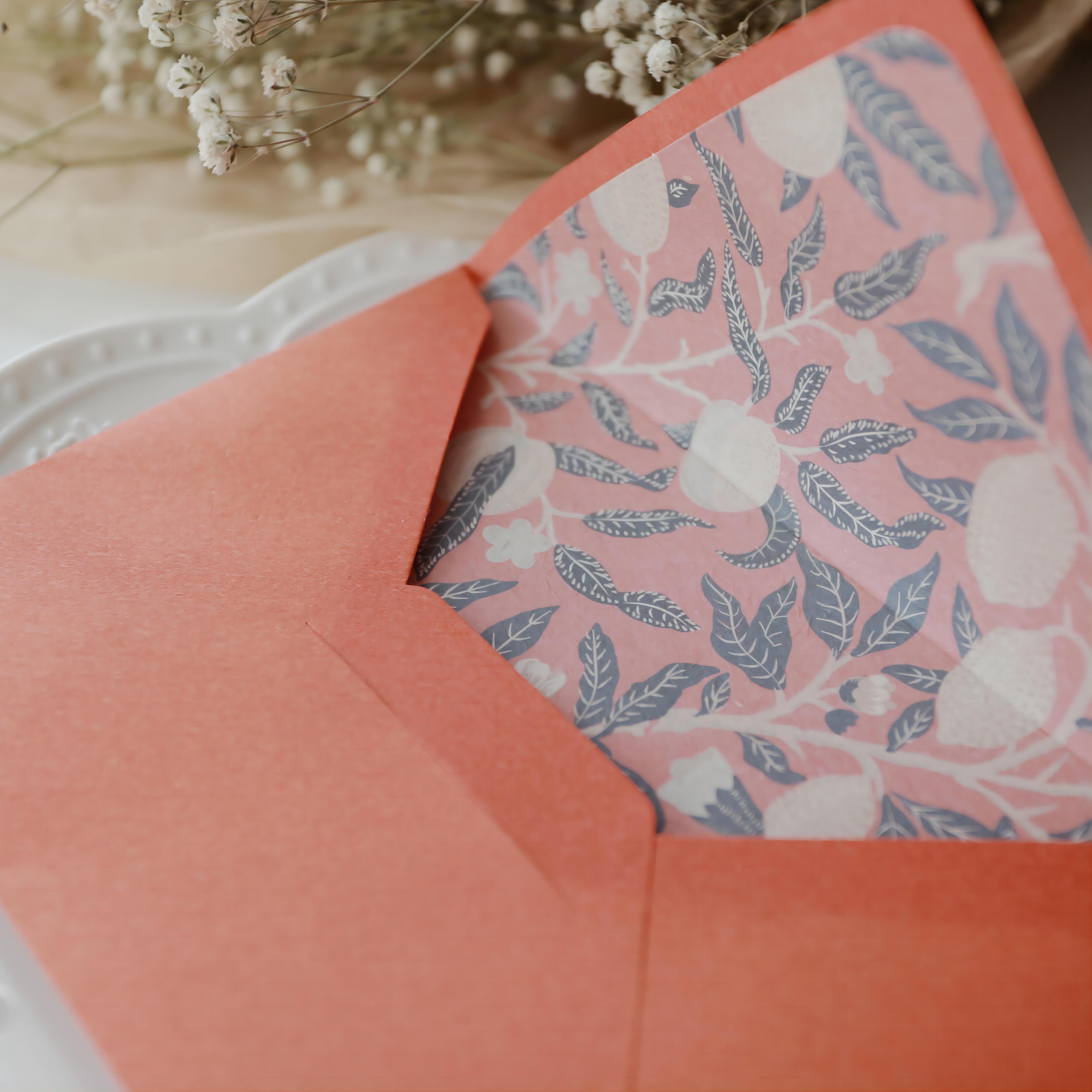 "Floral Elegance" vintage artistic envelope in a coral orange color, featuring an intricate floral and leaf pattern lining in soft pink and navy blue. The envelope is elegantly displayed on a white plate with a delicate embossed edge and surrounded by baby's breath flowers, creating an artistic and serene atmosphere. Perfect for special occasions or personal correspondence. Made from eco-friendly materials.