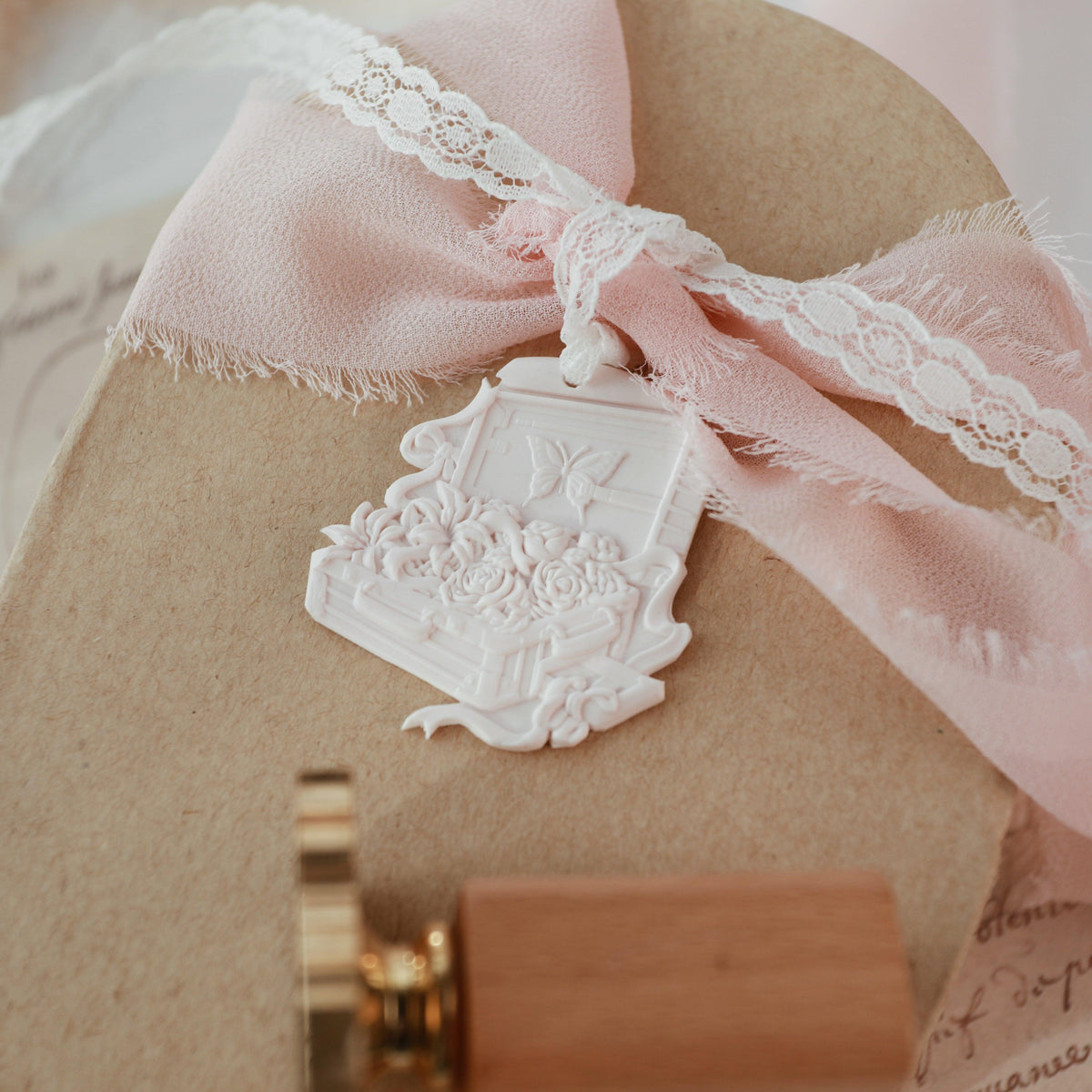 "Flower Journey" wax seal stamp showcasing a detailed floral design with roses and a butterfly on an open suitcase. The wax seal is crafted in a soft, ivory color and elegantly tied to a natural kraft box with a delicate pink ribbon and lace bow. A gold and wooden handle stamp is partially visible at the bottom, adding a touch of vintage charm. This setup, with its intricate details and soft color palette, is ideal for adding a romantic and elegant touch to gift packaging or correspondence.