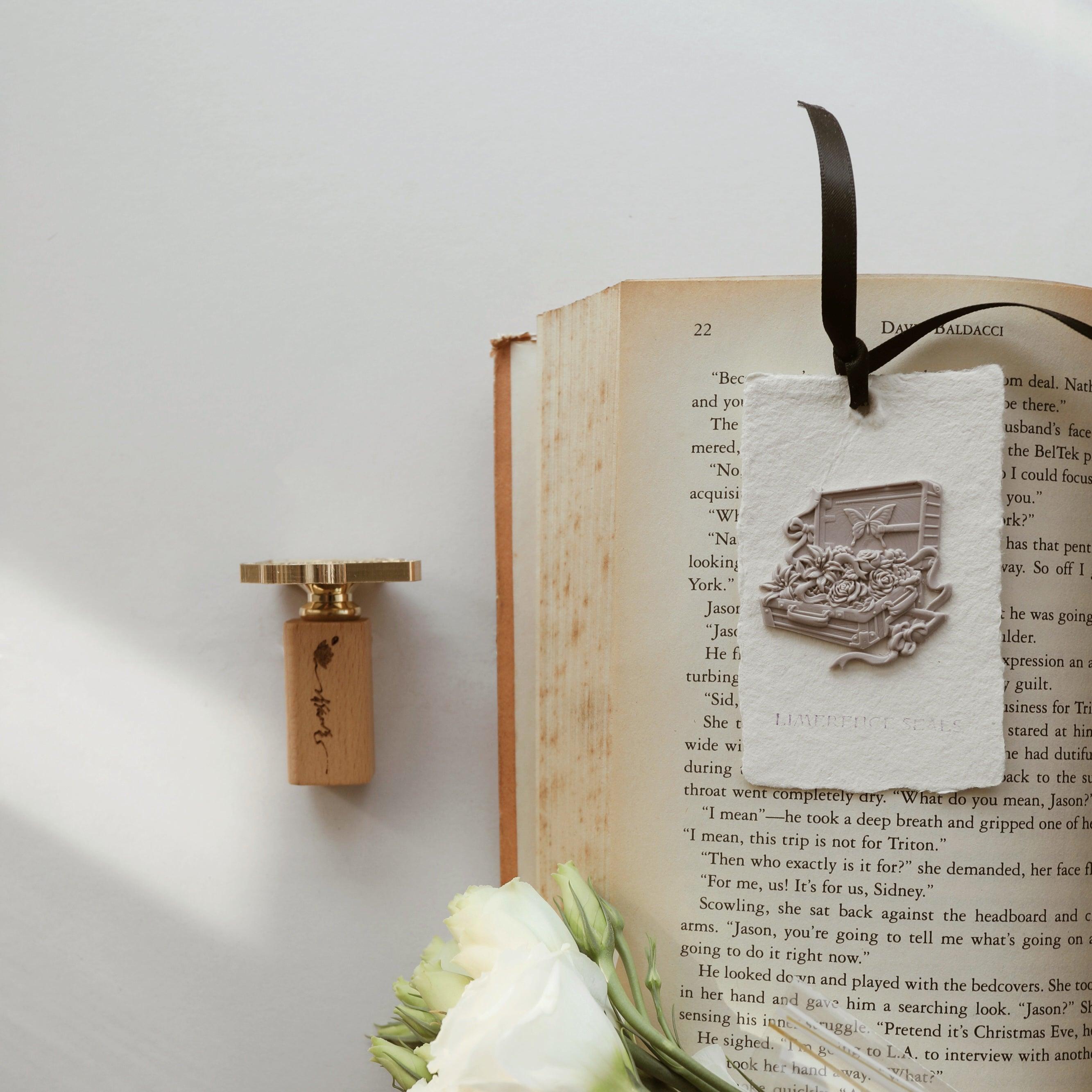 "Flower Journey" wax seal stamp featuring an intricate design of an open suitcase filled with roses and a butterfly. The wax seal is attached to a textured tag made from handmade paper, embossed with "LIMERENCE SEALS," and used as a bookmark in an open book with a black ribbon tassel. A wooden-handled wax stamp with a gold head rests nearby, alongside a bouquet of white roses, adding elegance to the scene. This setup blends literary charm with vintage elegance, perfect for book lovers and gift packaging.