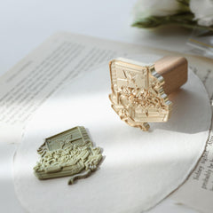 "Flower Journey" wax seal stamp displaying an intricate design of a suitcase with roses and a butterfly. The stamp, crafted from gold metal with a wooden handle, is placed on a textured paper along with a green wax imprint of the same design. An open book and a bouquet of white roses add a literary and elegant touch to the scene, ideal for enhancing gifts or correspondence with vintage charm.