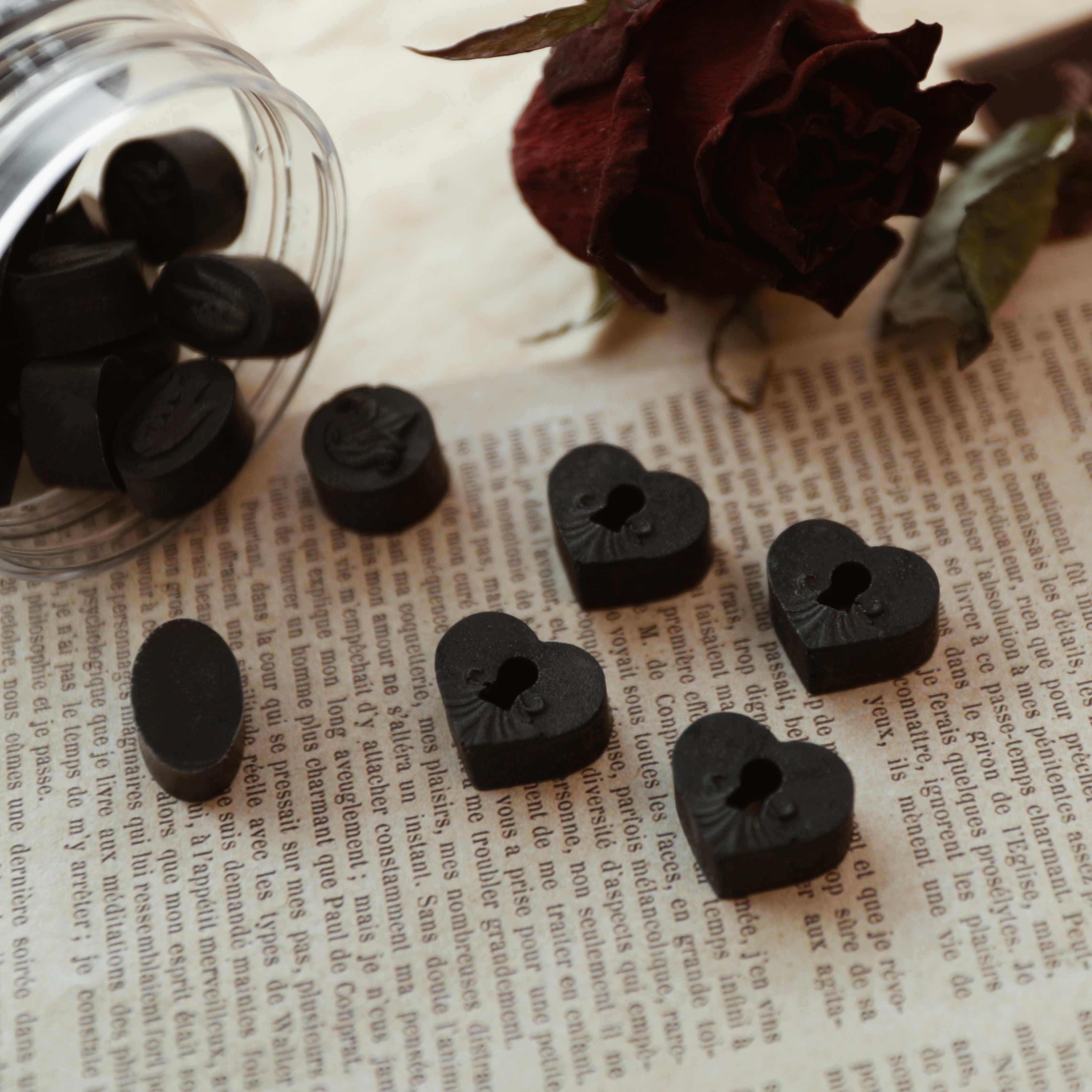 Black sealing wax beads from LimerenceSeals, featuring oval and heart shapes with intricate relief designs, scattered on a vintage-style French book page. A dried dark red rose enhances the mysterious and romantic atmosphere, ideal for letter sealing and decoration.