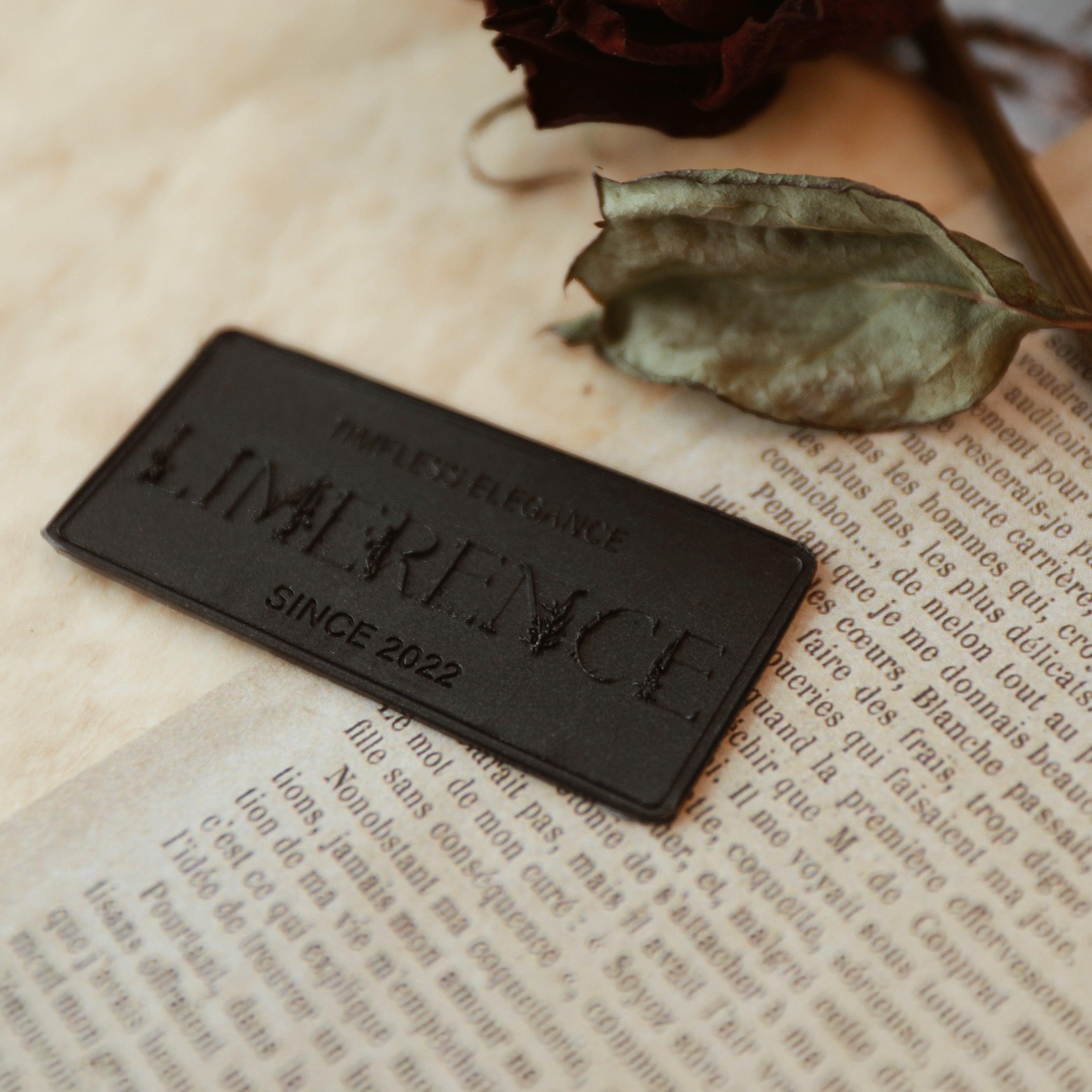 A black wax plaque engraved with 'LimerenceSeals Timeless Elegance Since 2022' on a vintage-style French book page. Accompanied by a dried dark red rose leaf, the scene creates an elegant and mysterious atmosphere, perfect for decoration and enhancing brand image.