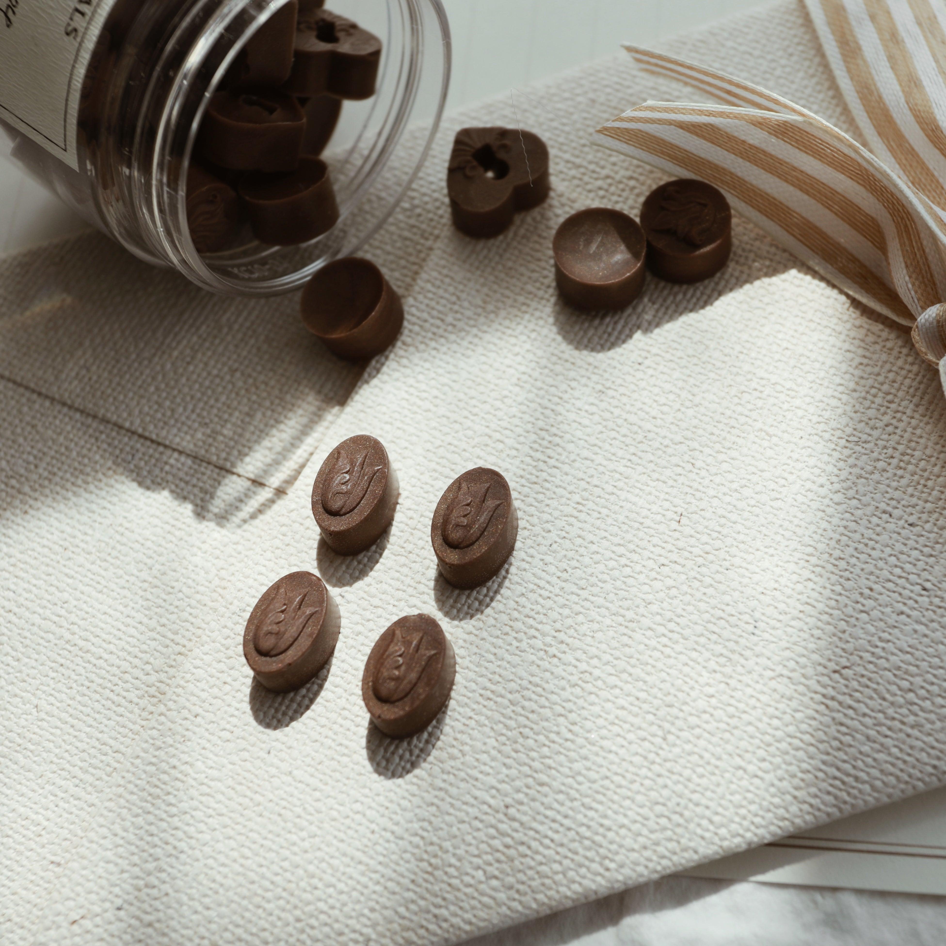 LimerenceSeals 'Late Autumn' handmade brown sealing wax beads scattered from a jar onto white fabric. The wax beads feature intricate relief designs in oval and heart shapes. A striped ribbon in the background adds a touch of warmth and elegance, perfect for letter sealing and crafting.