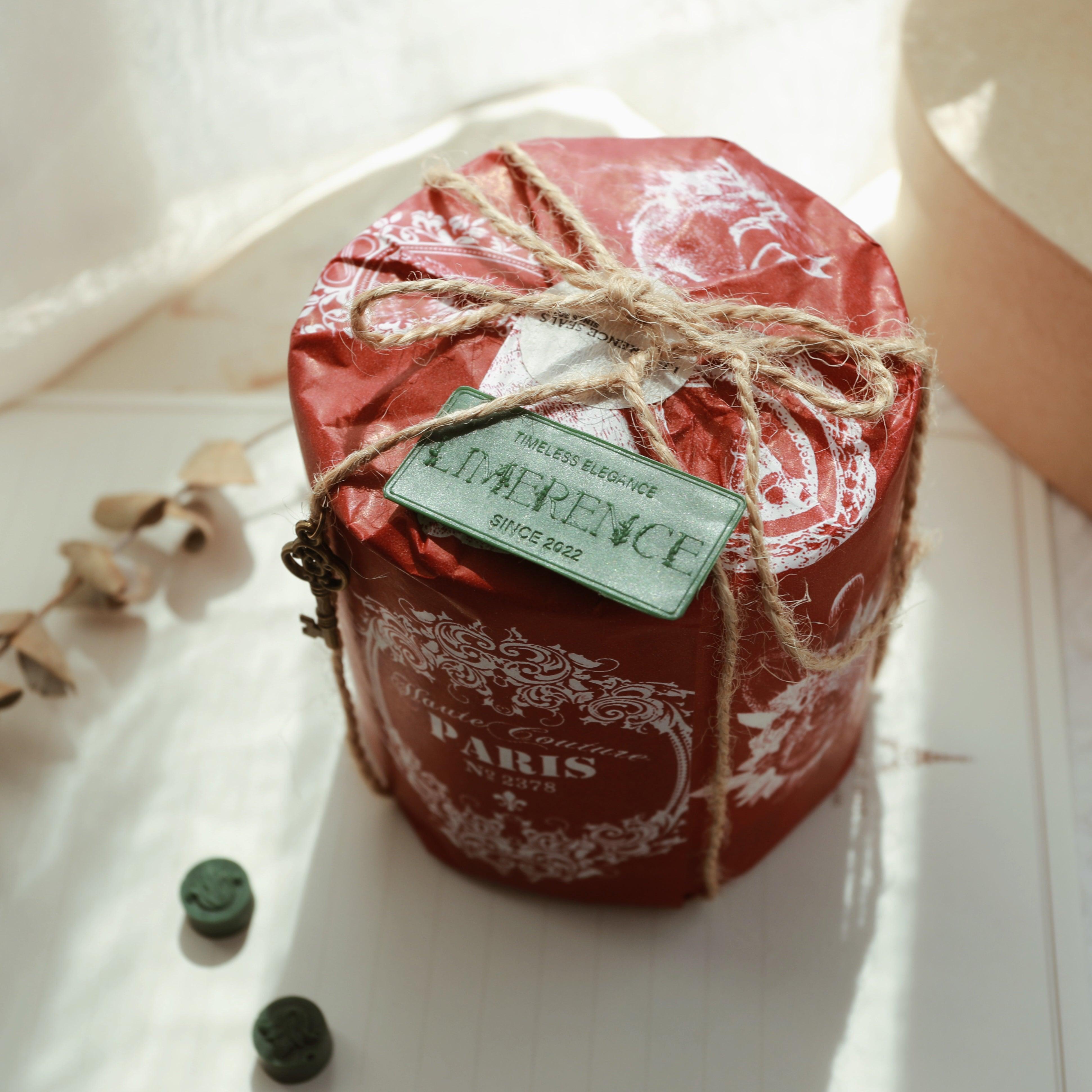 A gift wrapped in red paper with a twine bow and a green wax plaque engraved with 'LimerenceSeals Timeless Elegance Since 2022.' The scene includes dark green sealing wax beads and a dried eucalyptus branch, creating a classic and elegant atmosphere in soft natural light.