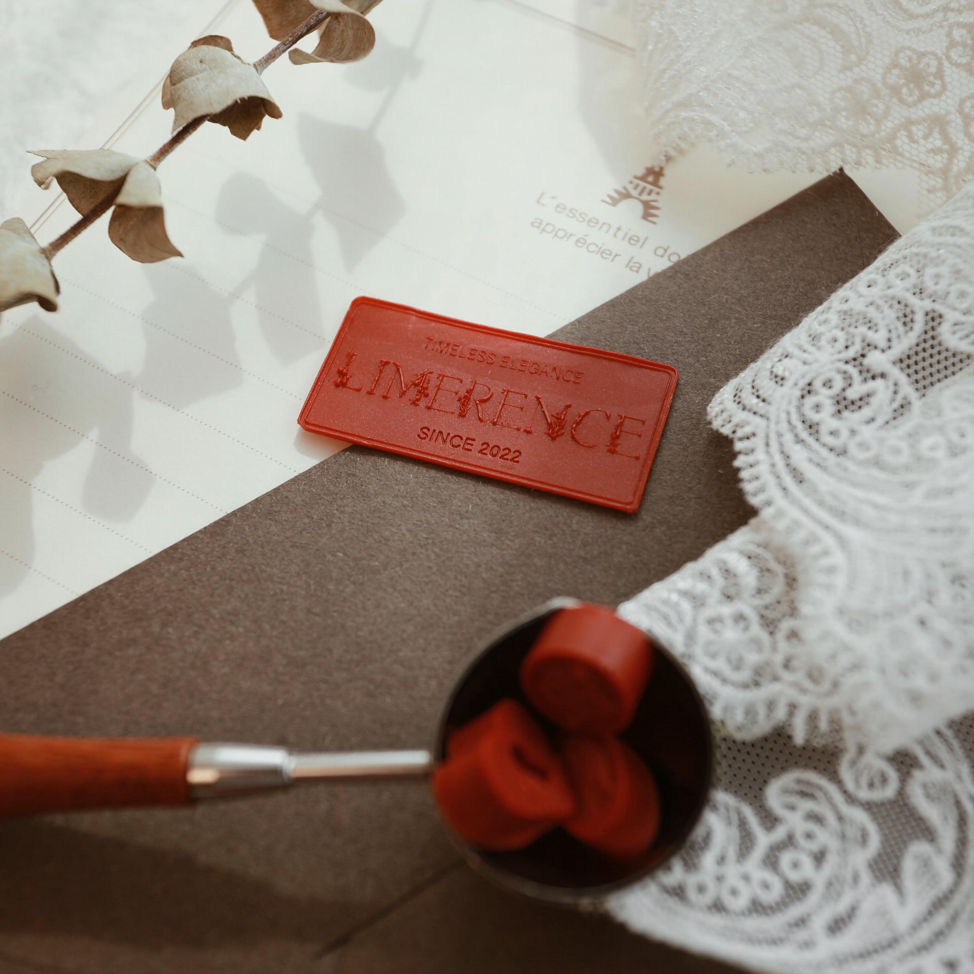 LimerenceSeals 'Late Autumn' handmade red sealing wax beads in a metal spoon, accompanied by a red wax plaque engraved with 'LimerenceSeals Since 2022.' The background features a brown envelope, white lace, and a dried eucalyptus branch, creating a vintage and elegant aesthetic.