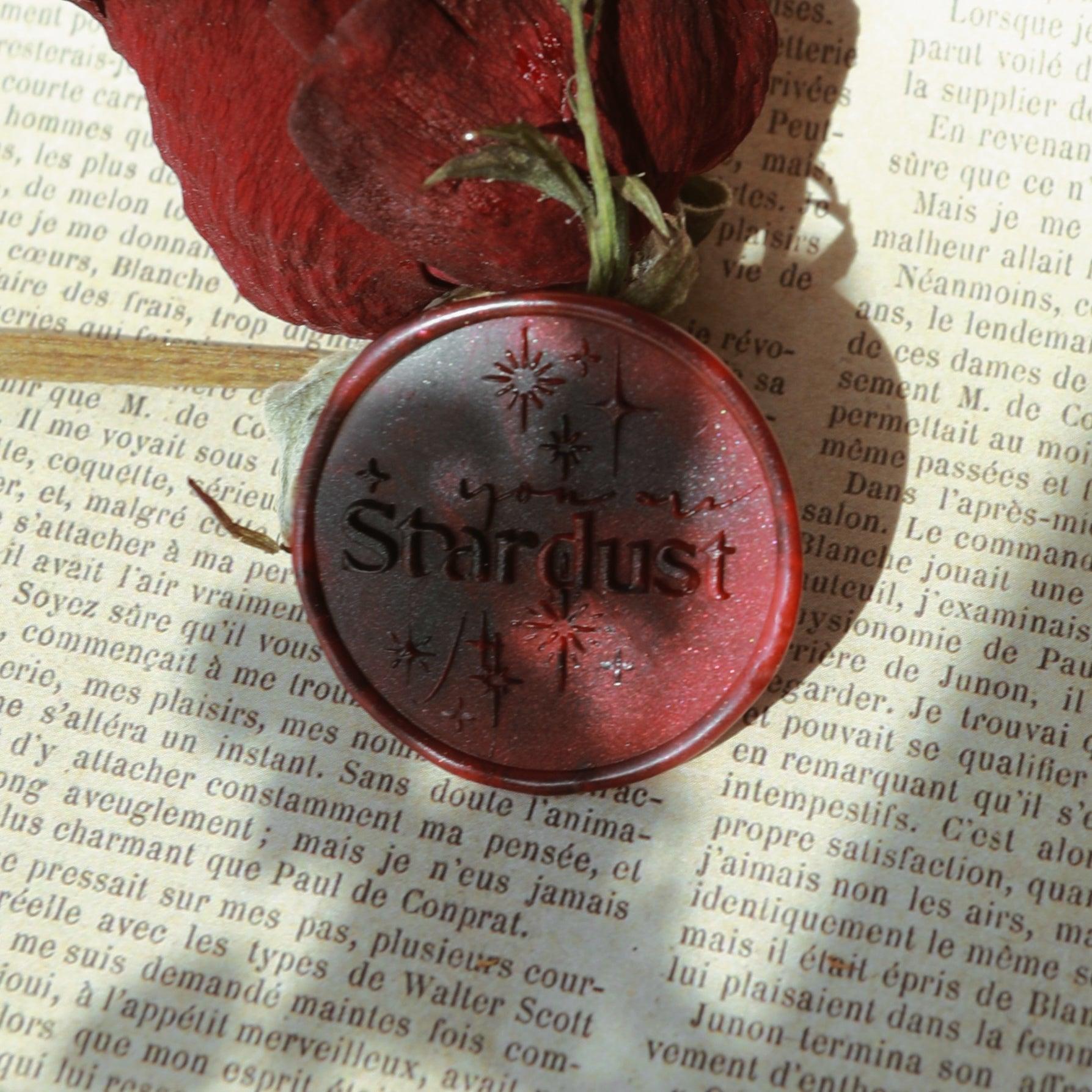 LimerenceSeals 'Stardust' red wax seal with engraved 'Stardust' text and star designs, set against an old French book page. Accompanied by red dried petals and green leaves, creating a vintage and romantic aesthetic.