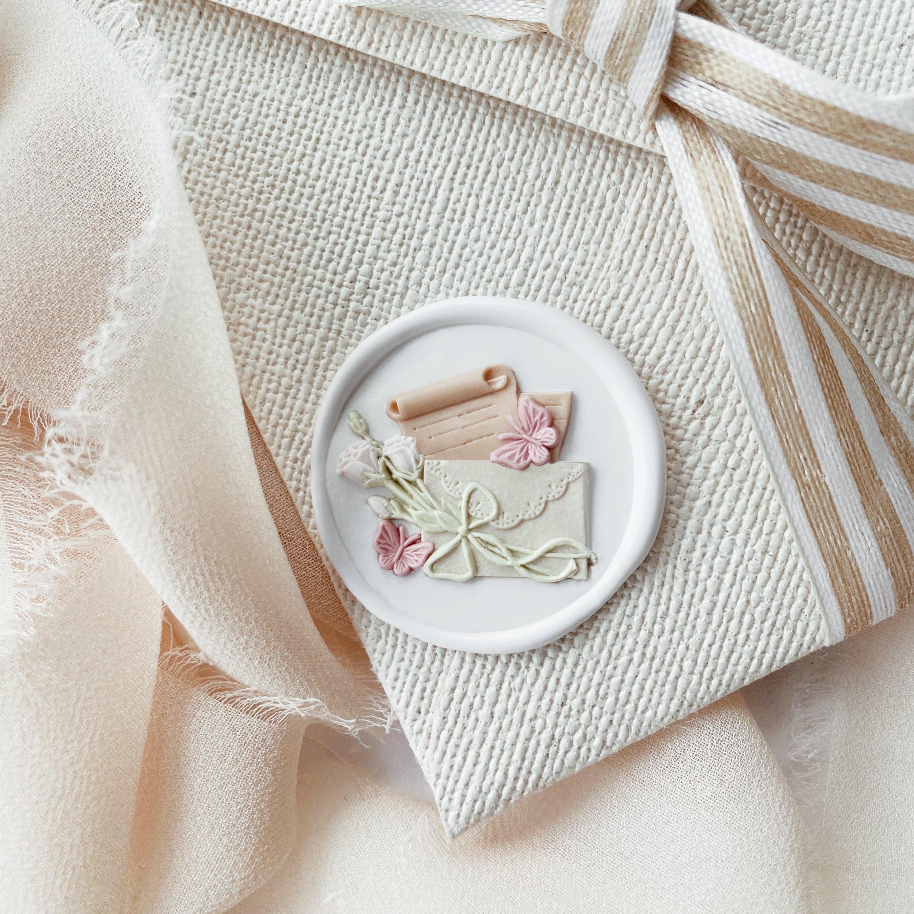 "Letters from Afar" wax seal featuring an intricate design of an envelope, scroll, and flowers. The seal is pressed into soft wax, elegantly placed on textured fabric with a striped ribbon. The delicate floral and ribbon details enhance the charm and sophistication of the setup, making it ideal for personalizing letters or special gifts.