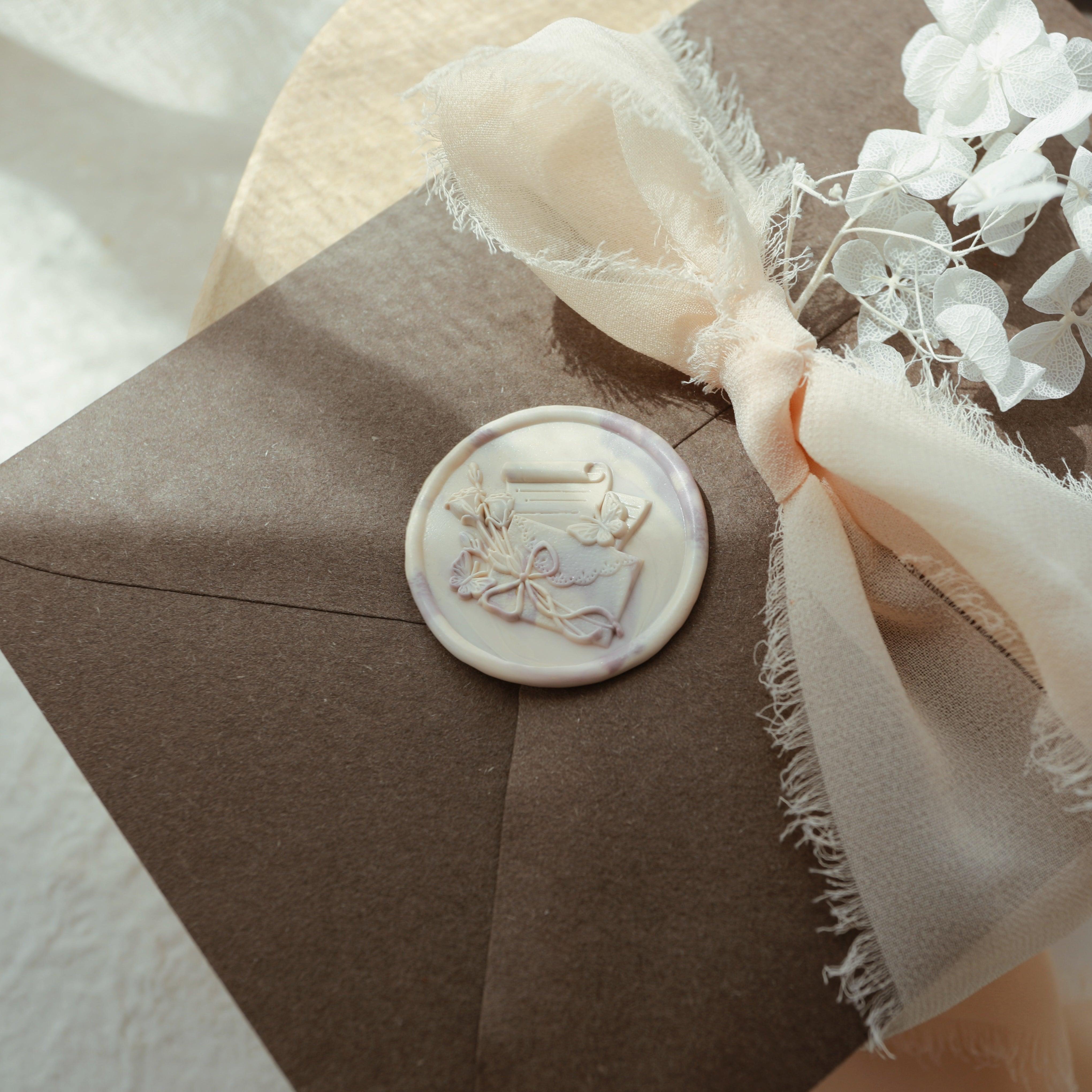 "Letters from Afar" wax seal featuring an elegant design of an envelope, scroll, and flowers. The seal is pressed into a marbled wax and placed on a dark brown envelope. It is adorned with a delicate peach-colored ribbon and white flowers, creating a sophisticated and artistic presentation. This setup is ideal for adding a personalized touch to invitations or special gifts.