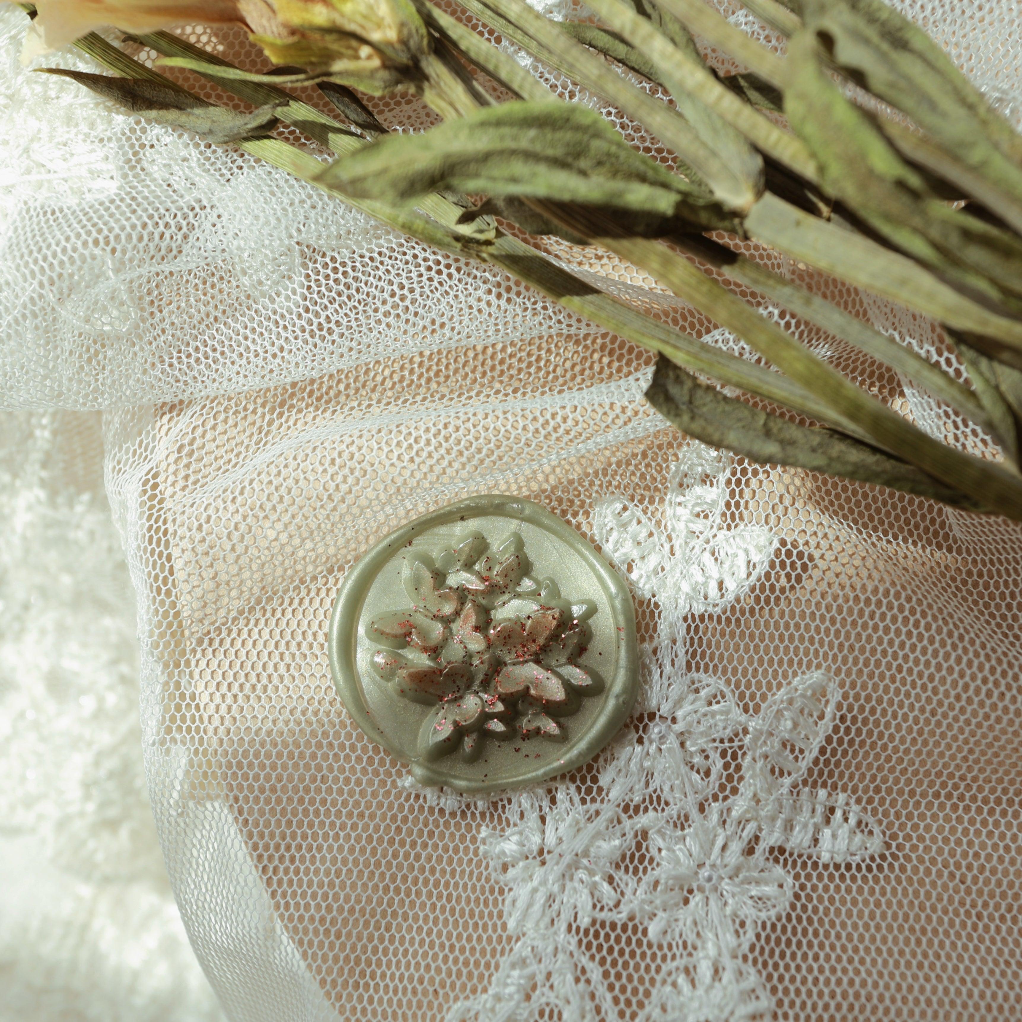 A jade green wax seal with intricate floral relief design and metallic specks, placed on lace fabric. The background features dried flowers, combining the wax seal's delicate design with the elegance of lace, creating a luxurious and romantic atmosphere, perfect for decorating wedding invitations or high-end letters.