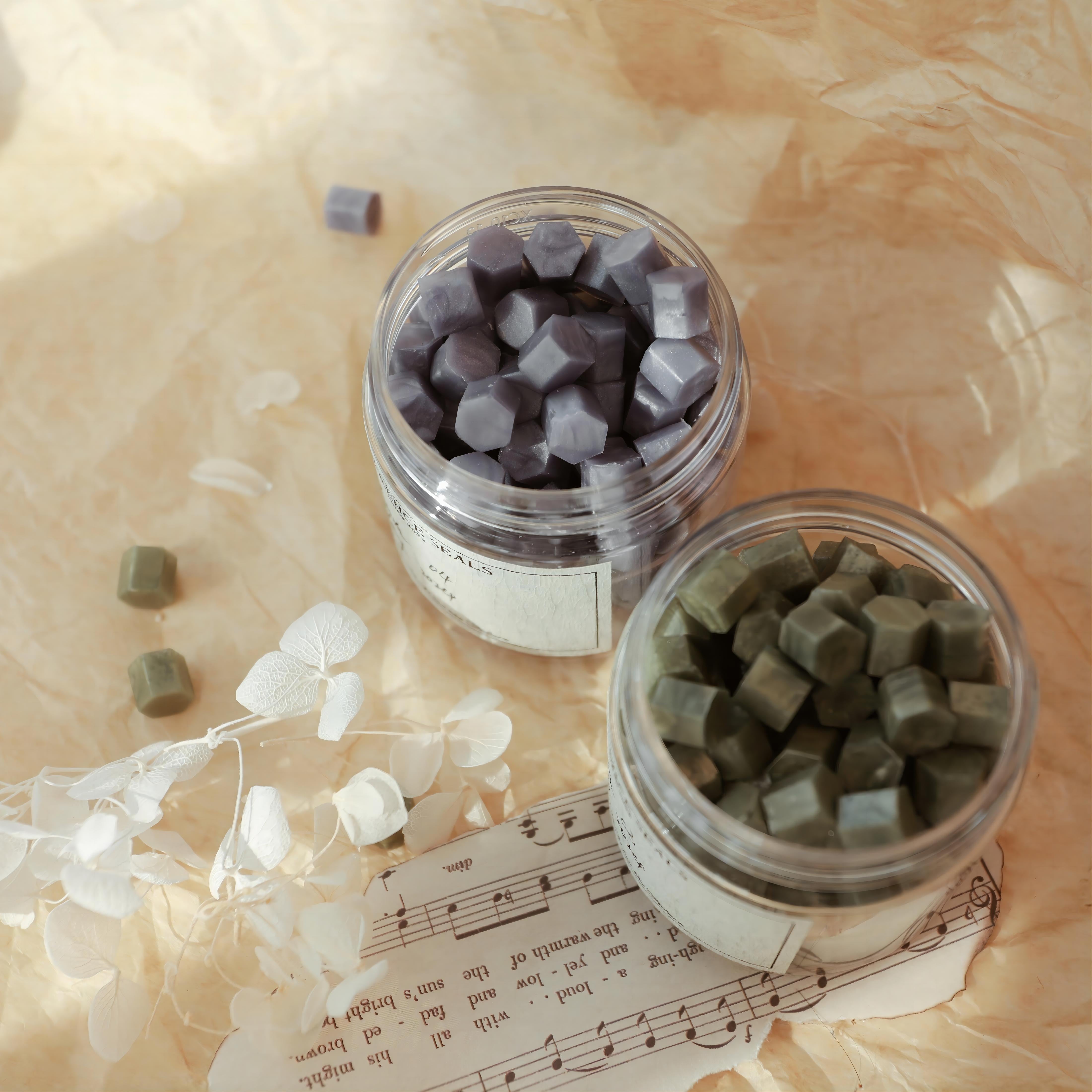 LimerenceSeals 'Lustr' handmade sealing wax beads in purple and green, contained in clear jars. The background features warm-toned paper, white dried flowers, and a vintage-style sheet music, creating a soft and elegant atmosphere. Perfect for letter sealing and crafts.