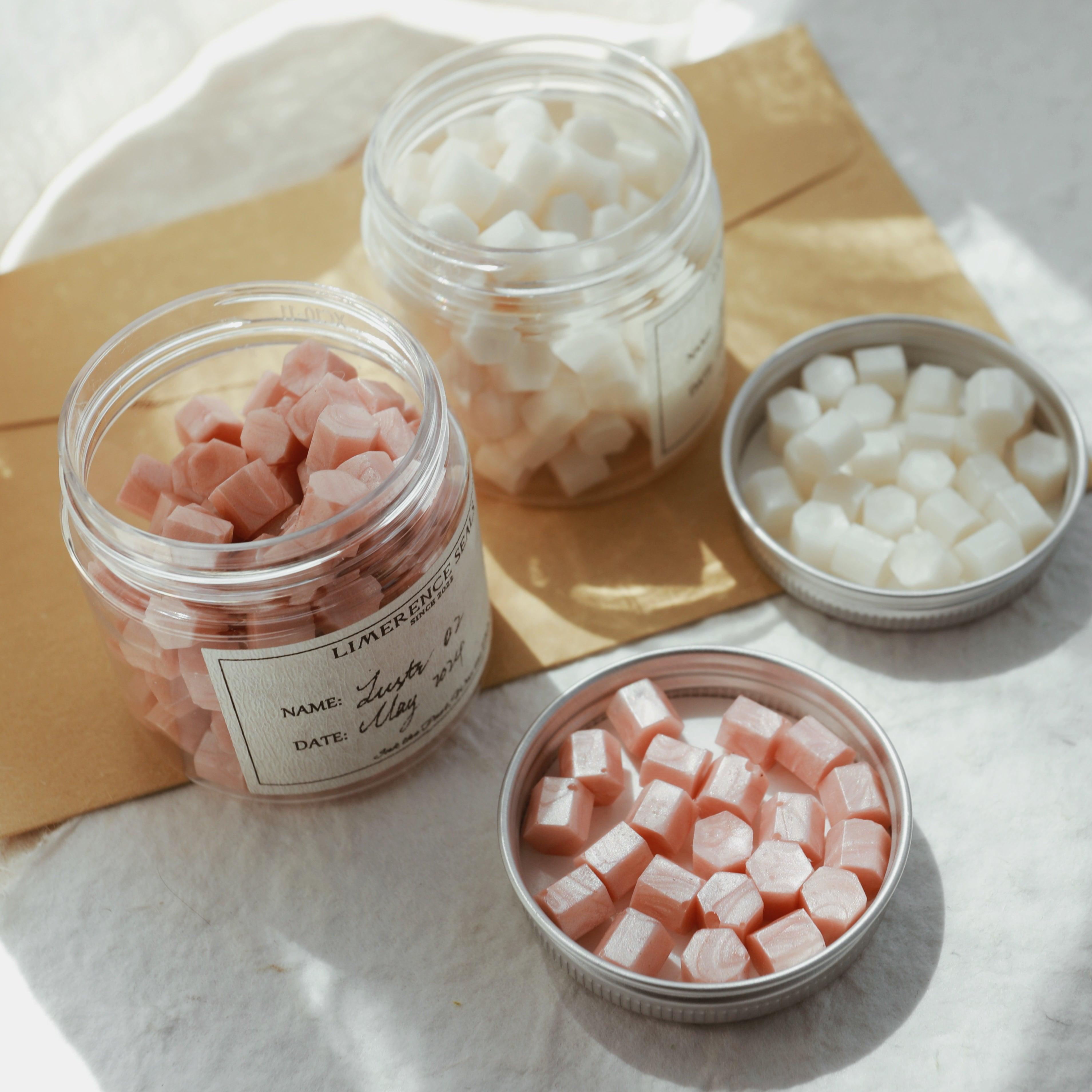 LimerenceSeals 'Lustr' handmade sealing wax beads in pink and white, contained in clear jars and jar lids. The background features a brown envelope, creating a bright and cozy atmosphere under soft natural light. Perfect for letter sealing and crafting.