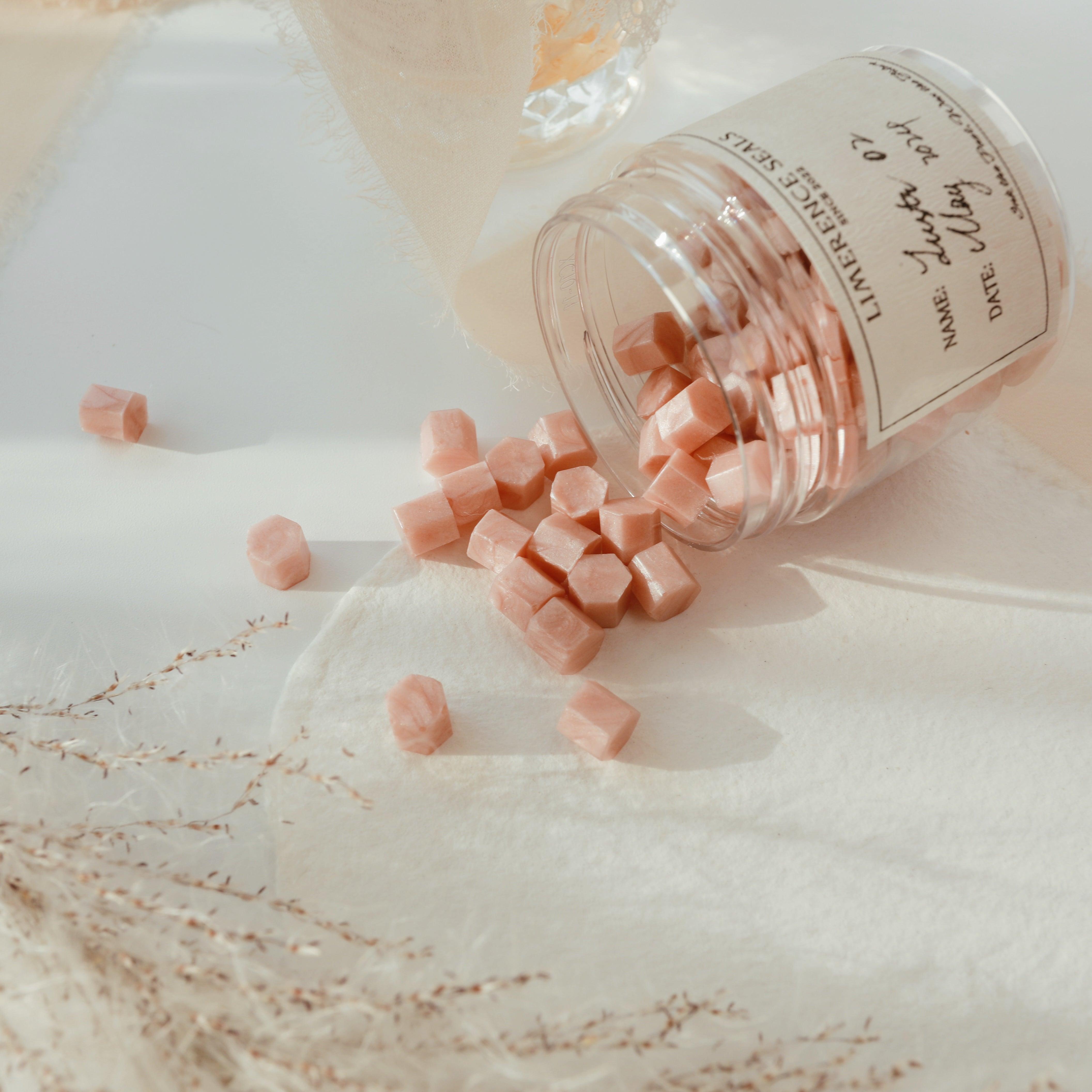 LimerenceSeals 'Lustr' handmade sealing wax beads in rose pink, spilling out from a clear jar onto a soft white background. The scene is enhanced by dried plant decorations and fabric, creating a romantic and cozy atmosphere. Perfect for letter sealing and crafts.