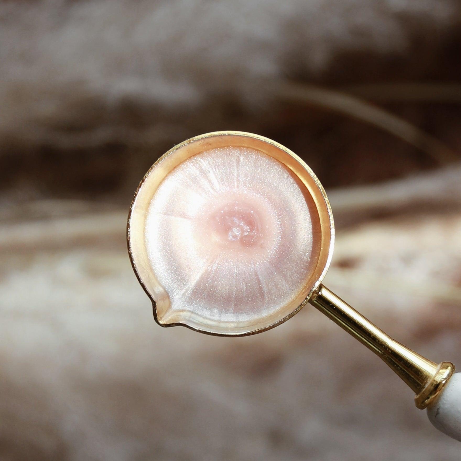 A gold spoon holding melted rose pink wax with a captivating pearlescent sheen, set against a soft fabric texture background. The image conveys an elegant and refined atmosphere, perfect for showcasing the process of making high-quality sealing wax.