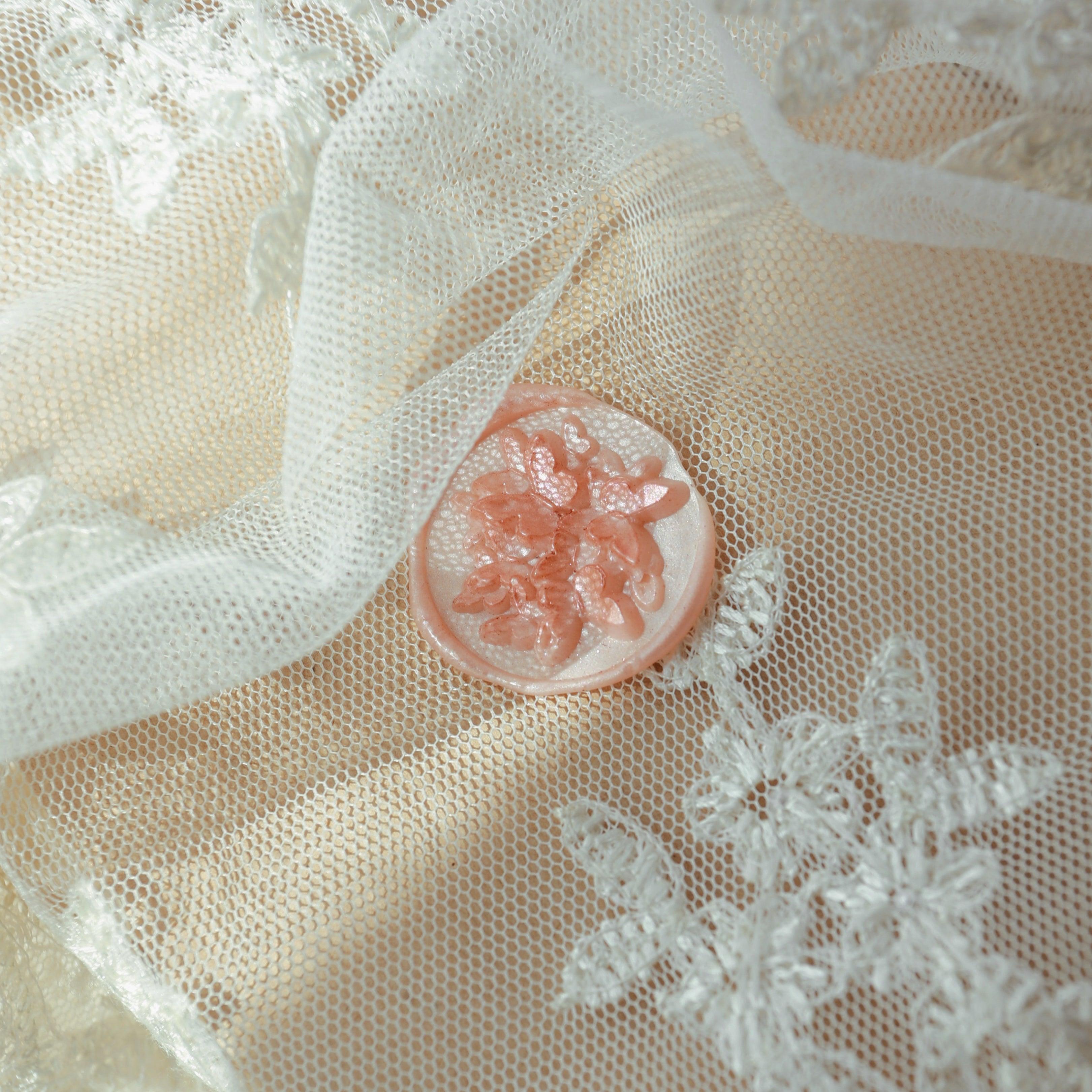 A rose pink wax seal with intricate floral relief design, placed on a lace fabric. The delicate texture of the lace complements the soft design of the wax seal, creating a romantic and elegant atmosphere, perfect for decorating wedding invitations or exquisite letters.