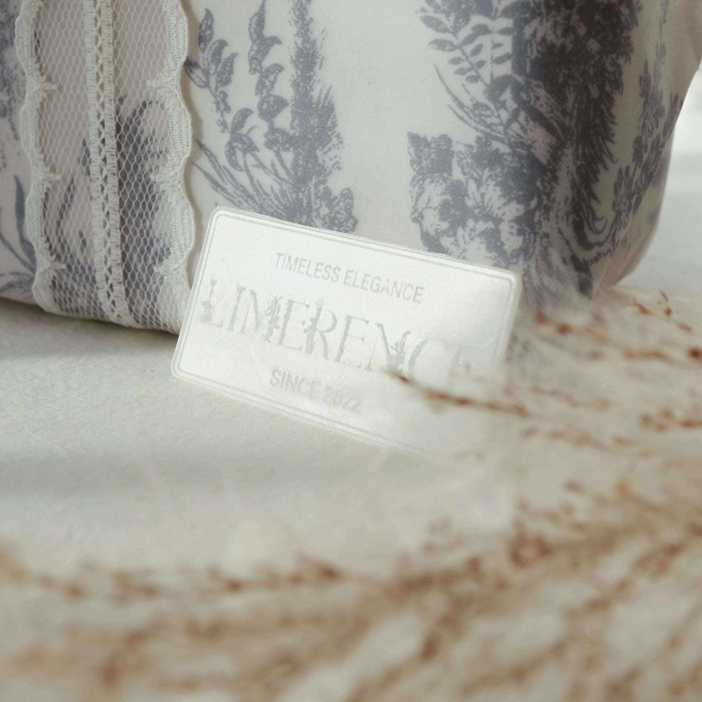 A white wax plaque engraved with 'LimerenceSeals Timeless Elegance Since 2022' placed on a white fabric. The background features fabric with gray floral patterns and dried plant decorations, creating an elegant and soft atmosphere, perfect for brand display and decoration.