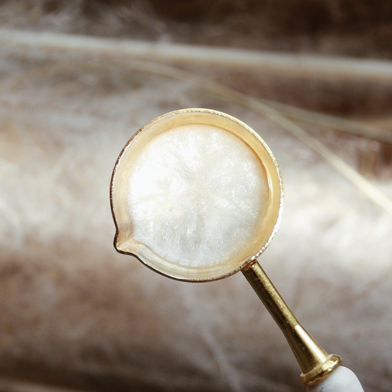 A gold spoon holding melted white wax with a pearlescent sheen, set against a soft fabric texture background. The scene creates an elegant and luxurious atmosphere, perfect for showcasing the high-quality sealing wax making process.