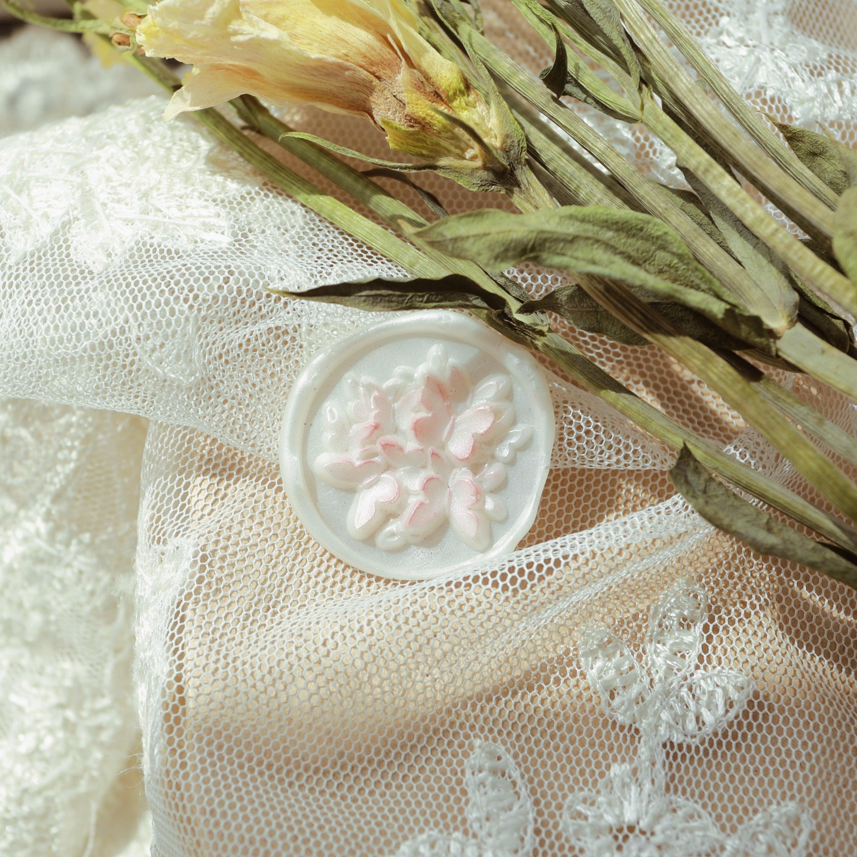 A white wax seal with intricate pink floral relief design, placed on lace fabric and dried yellow flowers. The delicate design of the wax seal complements the textured background, creating a romantic and elegant atmosphere, perfect for decorating wedding invitations or exquisite letters.