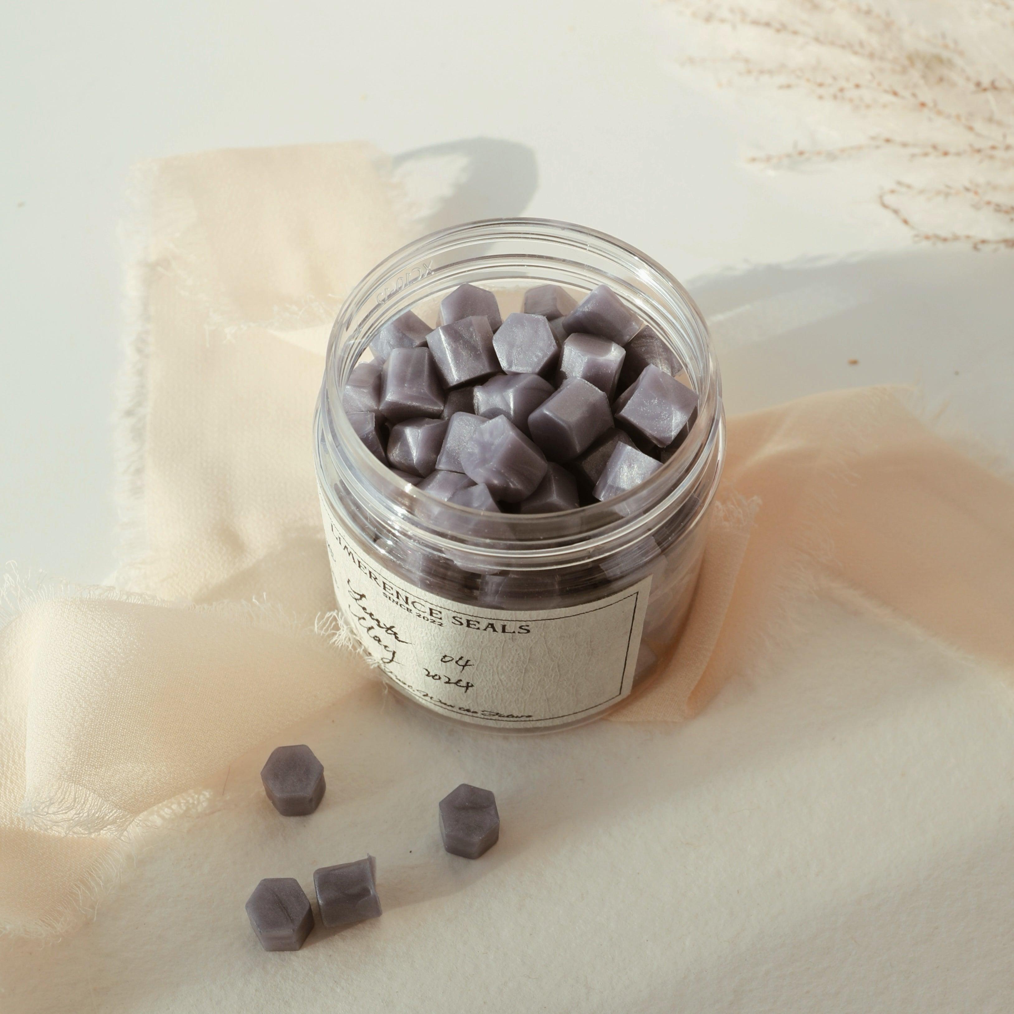 LimerenceSeals 'Lustr' handmade sealing wax beads in twilight blue, contained in a clear jar with a few beads scattered on soft beige fabric. The scene conveys a serene and elegant atmosphere, perfect for letter sealing and crafting.