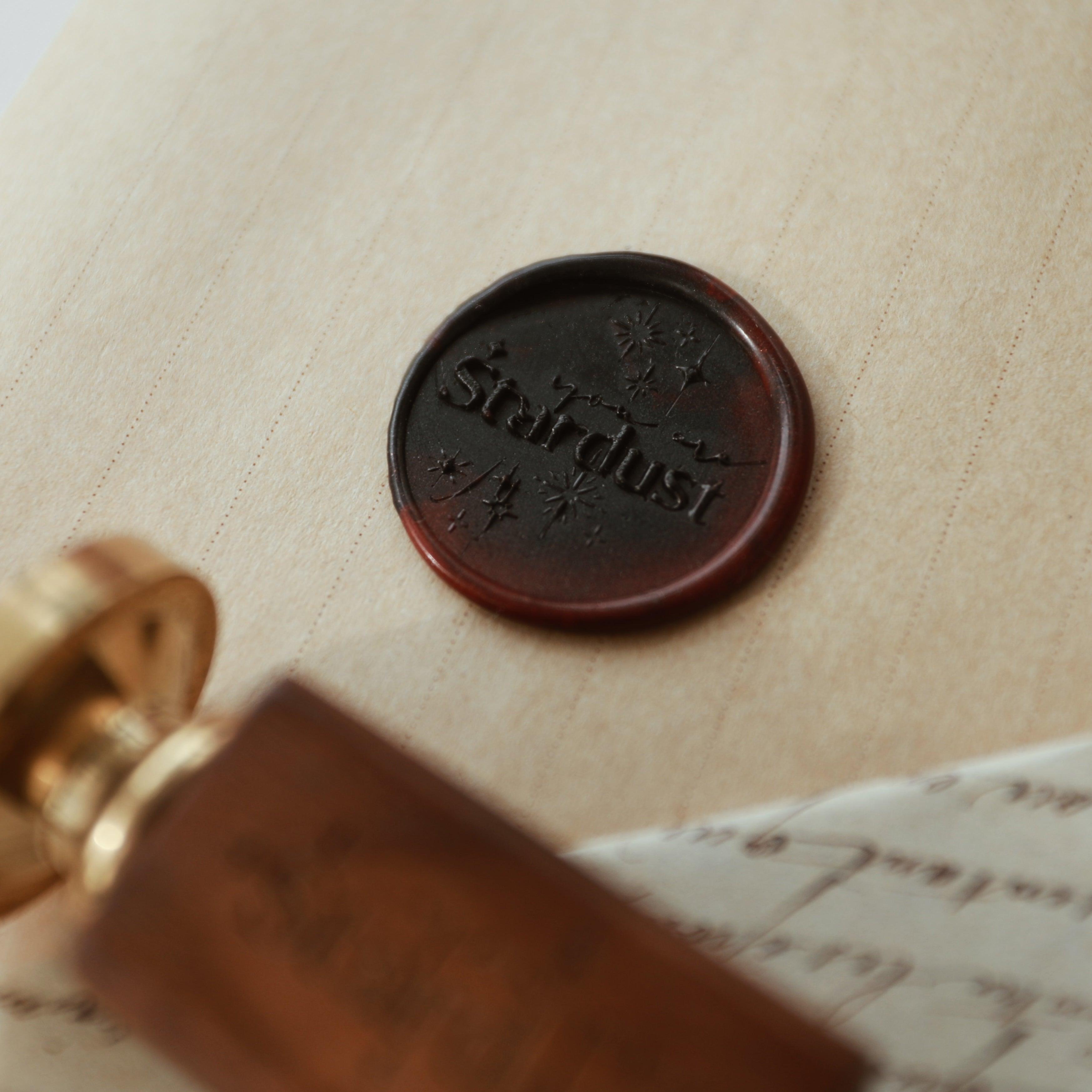 The "Ethereal Stardust" wax seal, featuring the word "Stardust" and delicate star motifs, is pressed into a rich dark wax on a vintage-style parchment. The seal is accompanied by a wooden-handled stamp with a gold head, partially visible in the background. This setup exudes a sense of elegance and mystery, perfect for adding a touch of celestial charm to letters and invitations.