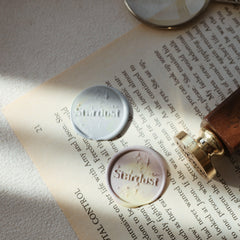 The "Ethereal Stardust" wax seals, featuring the word "Stardust" and star motifs, are pressed into marbled wax blends. They are placed on an open book, with a wooden-handled stamp nearby. A magnifying glass partially visible in the background adds a touch of curiosity and exploration. This setup beautifully blends celestial charm with a literary theme, perfect for enhancing letters, invitations, or book collections.