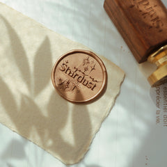 The "Ethereal Stardust" wax seal, featuring the phrase "you're Stardust" and star motifs, is pressed into a metallic wax on a piece of textured parchment. Nearby is a wooden-handled stamp with a gold head, adding elegance and charm to the scene. The setup is perfect for adding a celestial touch to letters, invitations, or special gifts.