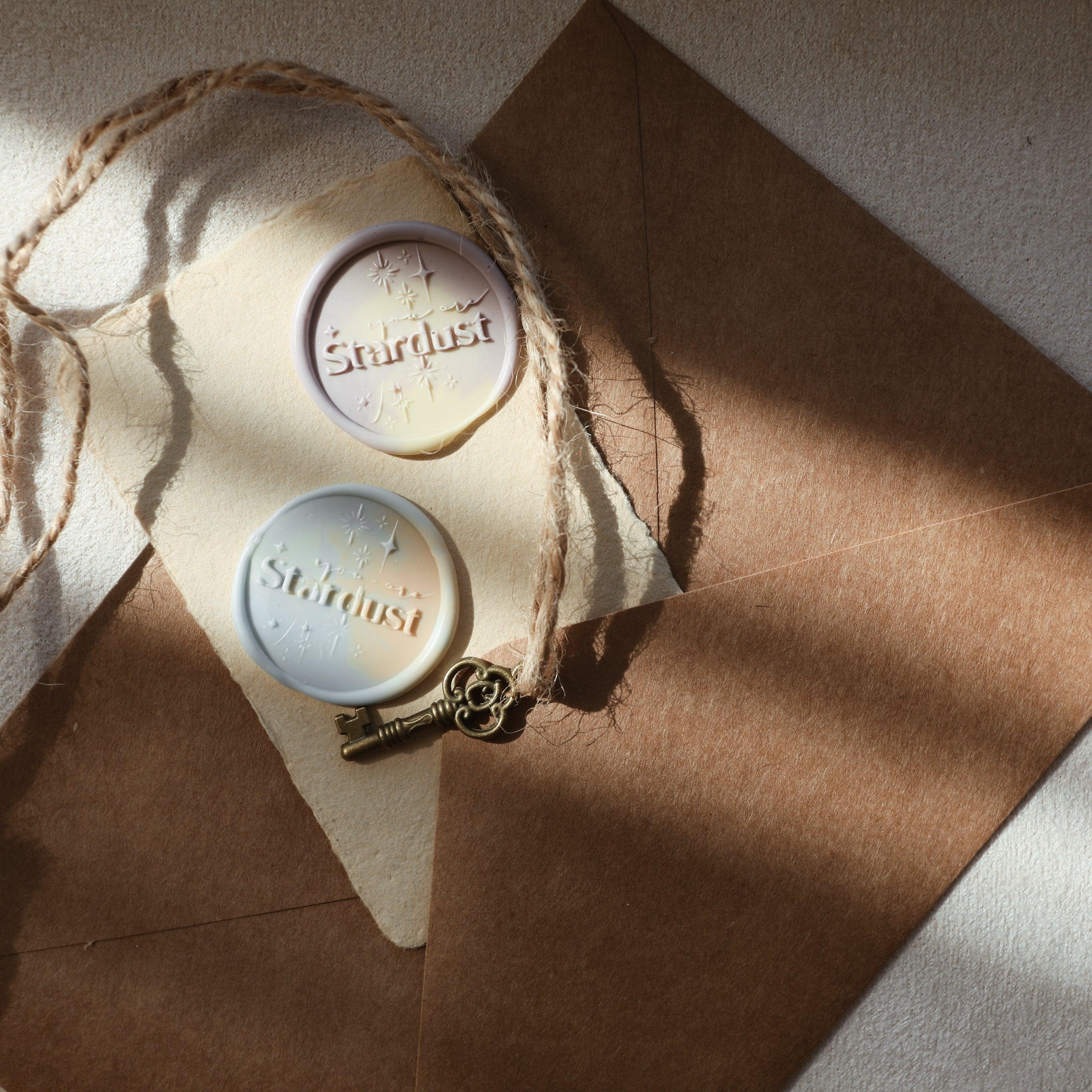 The "Ethereal Stardust" wax seals, featuring the word "Stardust" and star motifs, are pressed into marbled wax. The seals are placed on a piece of textured parchment atop a brown kraft envelope, along with a vintage-style key tied to a piece of twine. This setup exudes a sense of mystery and elegance, perfect for enhancing letters or invitations with a celestial touch.