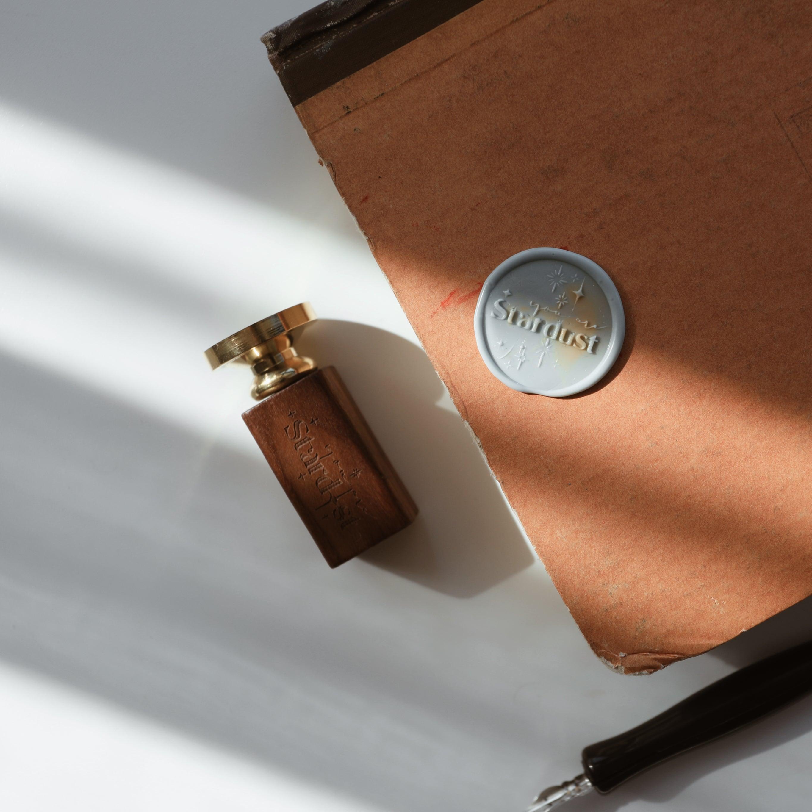 The "Ethereal Stardust" wax seal, featuring the word "Stardust" and star motifs, is pressed into marbled wax on a rustic brown book cover. A wooden-handled stamp with a gold head is placed nearby, along with a black wax seal pen. The setup exudes a sense of vintage elegance, ideal for adding a celestial touch to journals, letters, or creative projects.