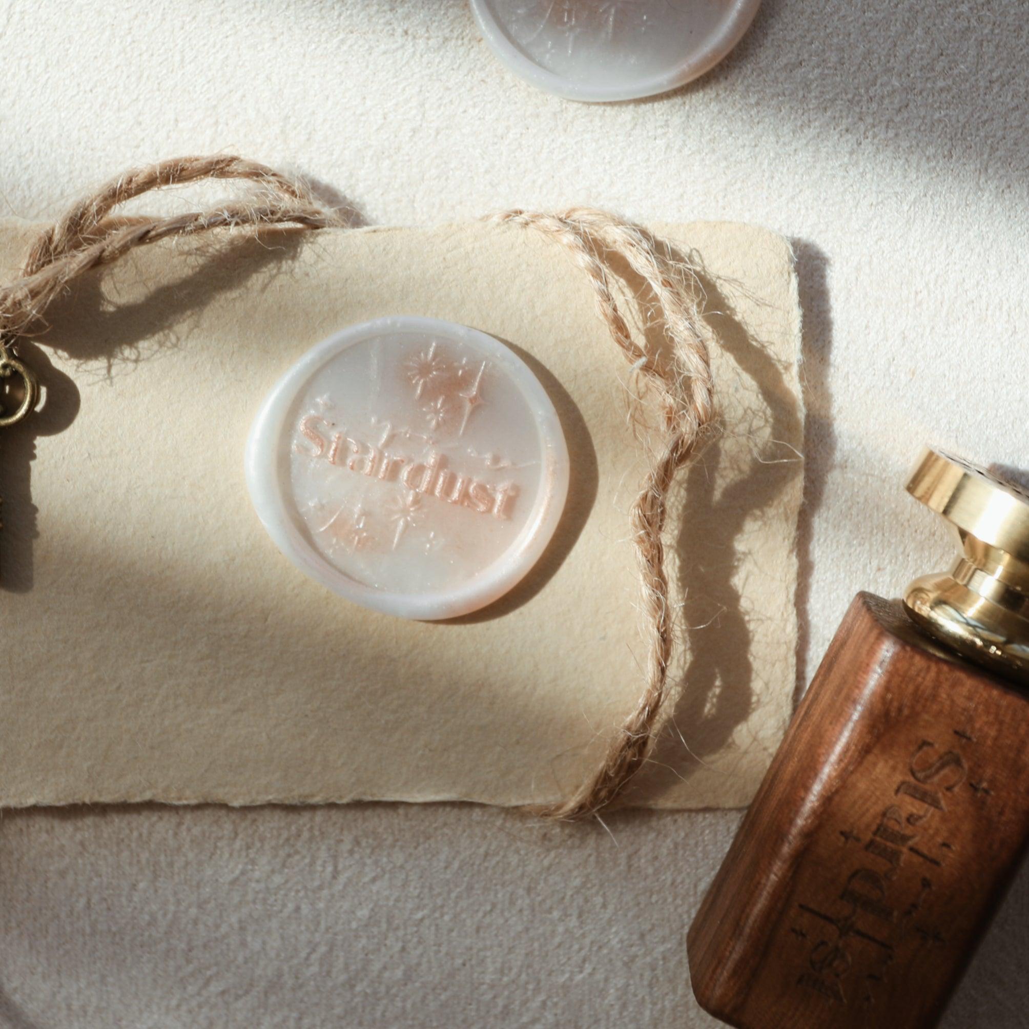 The "Ethereal Stardust" wax seal, embossed with the word "Stardust" and star motifs, is pressed into marbled wax. The seal sits on textured parchment, with a vintage-style key attached to twine nearby, and a wooden-handled stamp partially visible at the edge. The arrangement conveys a sense of elegance and mystery, perfect for embellishing letters and invitations with a celestial theme.