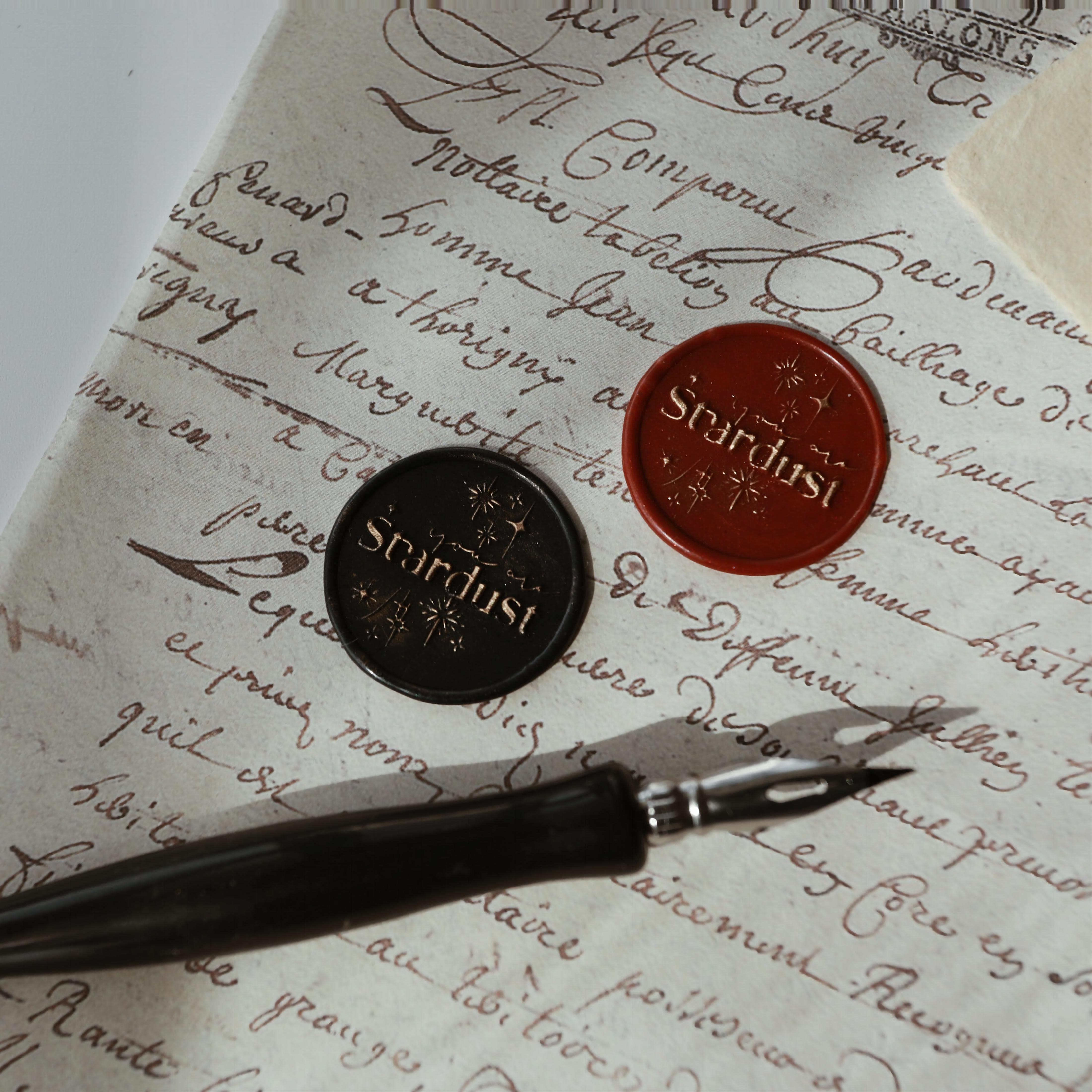 The "Ethereal Stardust" wax seals feature the word "Stardust" and star motifs, with one seal embossed in black wax with gold highlights and the other in red wax with gold highlights. They are placed on vintage-style parchment with handwritten script, accompanied by a black calligraphy pen. This arrangement exudes a sense of timeless elegance and mystique, perfect for enhancing letters, invitations, or creative projects with a celestial touch.