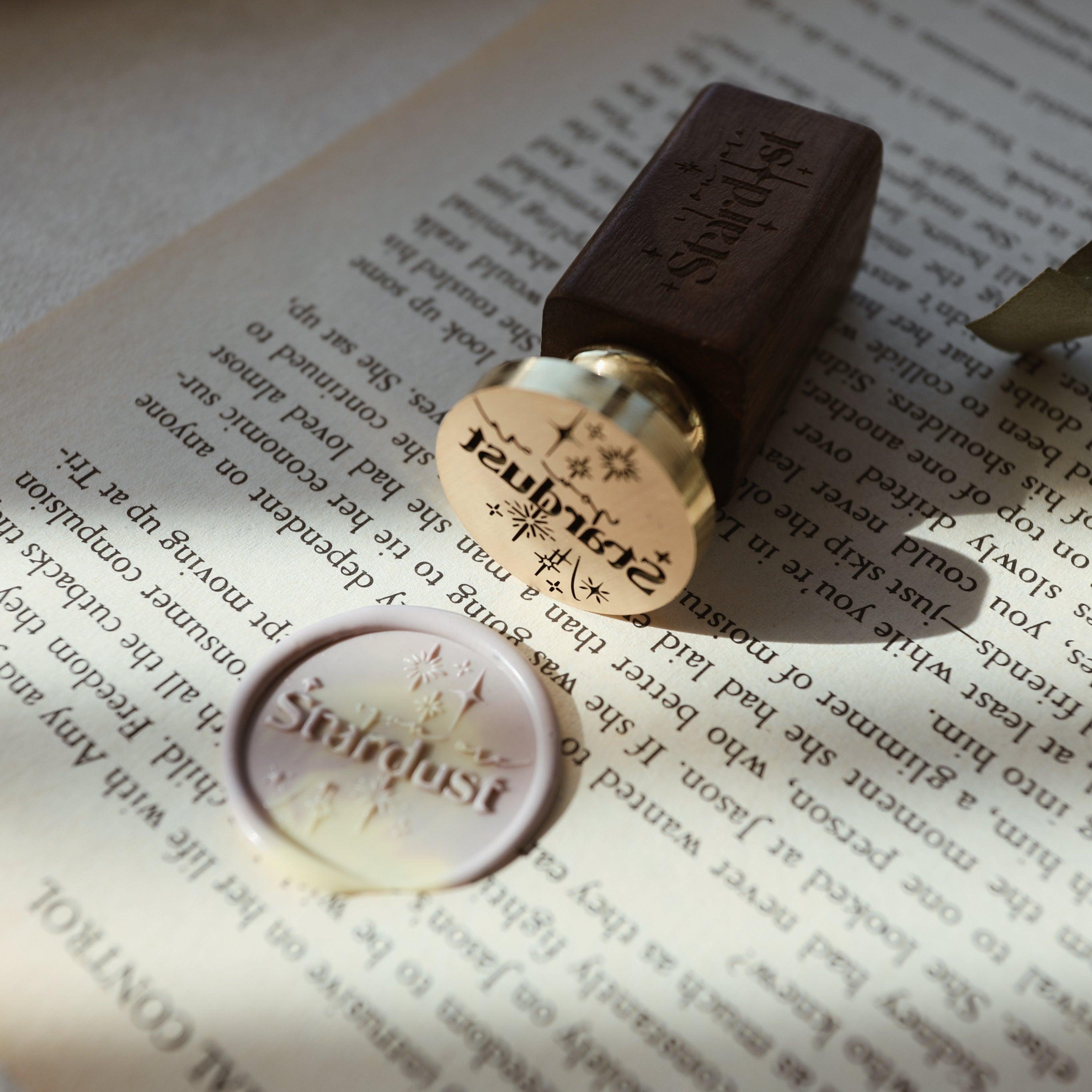 The "Ethereal Stardust" wax seal stamp, featuring the word "Stardust" and star motifs, is shown next to a wax seal embossed in marbled wax. The stamp has a gold head and a wooden handle, resting on an open book. The combination of the wax seal and the stamp creates a celestial and elegant theme, perfect for adding a touch of magic to letters, invitations, or journals.