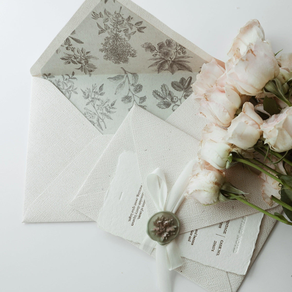 A beautifully arranged flat lay featuring Limerence Seals' "Textures of Time" artistic envelope, with an intricate floral lining and a card secured by a white ribbon and a floral sealing wax stamp. The elegant design is complemented by a bouquet of soft pink roses, creating a harmonious and timeless aesthetic that captures the essence of thoughtful correspondence and refined elegance.