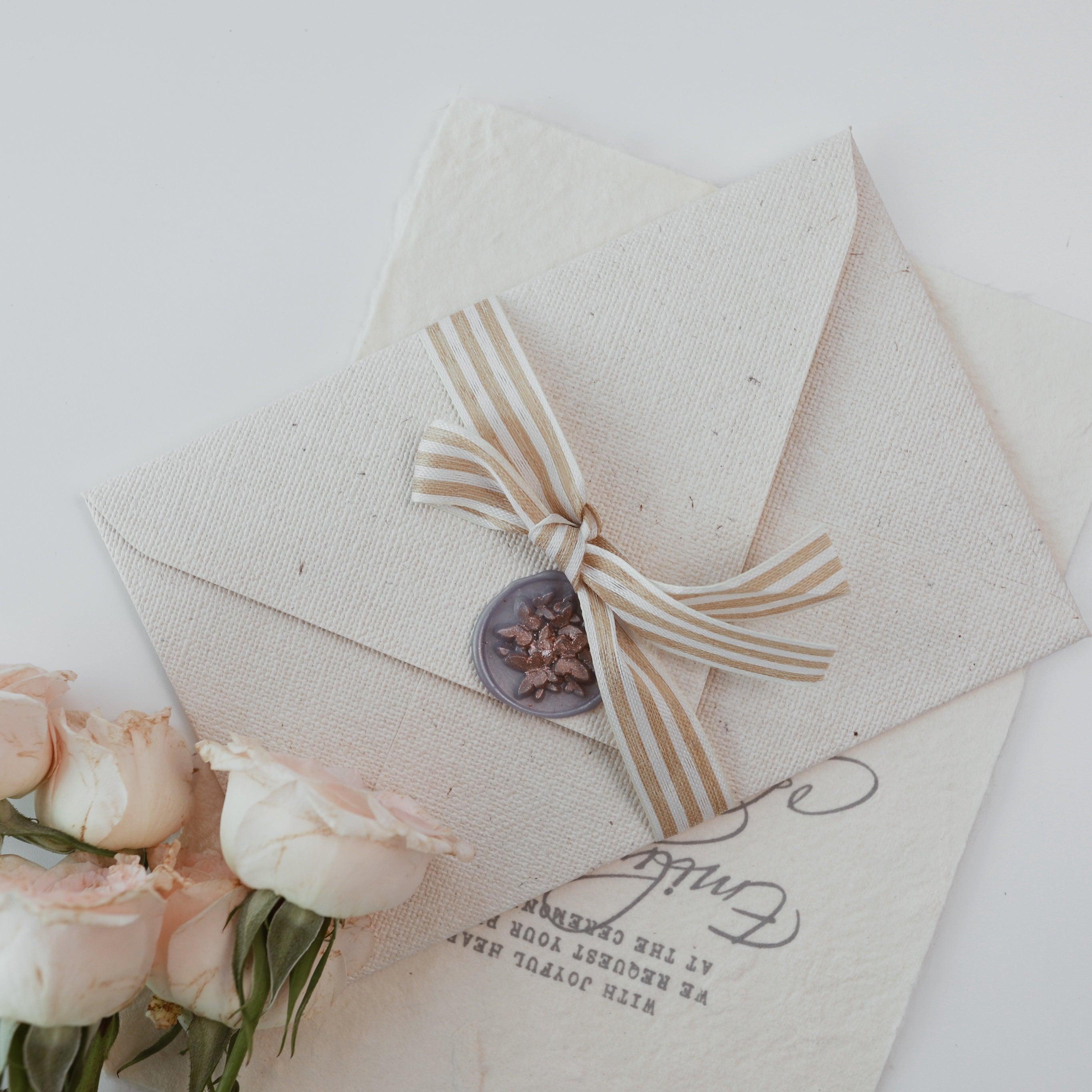 A beautifully textured beige envelope from Limerence Seals' "Textures of Time" collection, elegantly sealed with a striped ribbon and a floral wax seal. The envelope is set against an aged, handwritten letter and paired with a bouquet of soft pink roses, creating a timeless and sophisticated aesthetic perfect for special occasions and heartfelt messages.