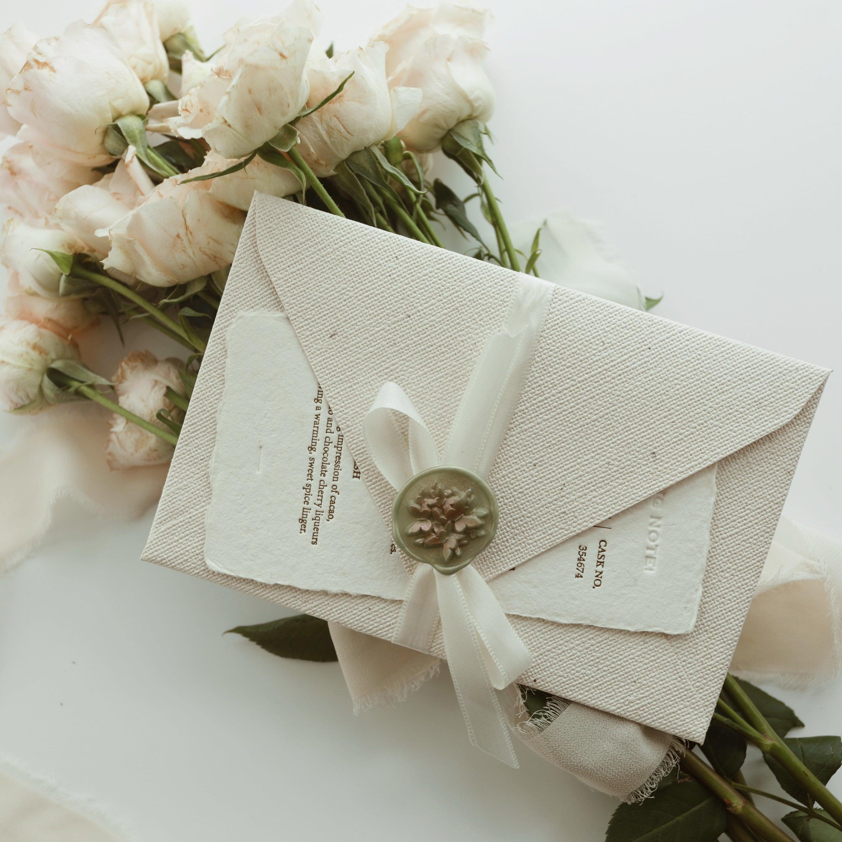 The Limerence Seals "Textures of Time" envelope, crafted from a sophisticated textured material, is elegantly sealed with a delicate ivory ribbon and a floral wax seal in soft green. The envelope rests atop an ornate vintage paper with intricate typography, surrounded by a bouquet of light pink roses, creating a romantic and elegant scene perfect for conveying heartfelt messages and special occasions.