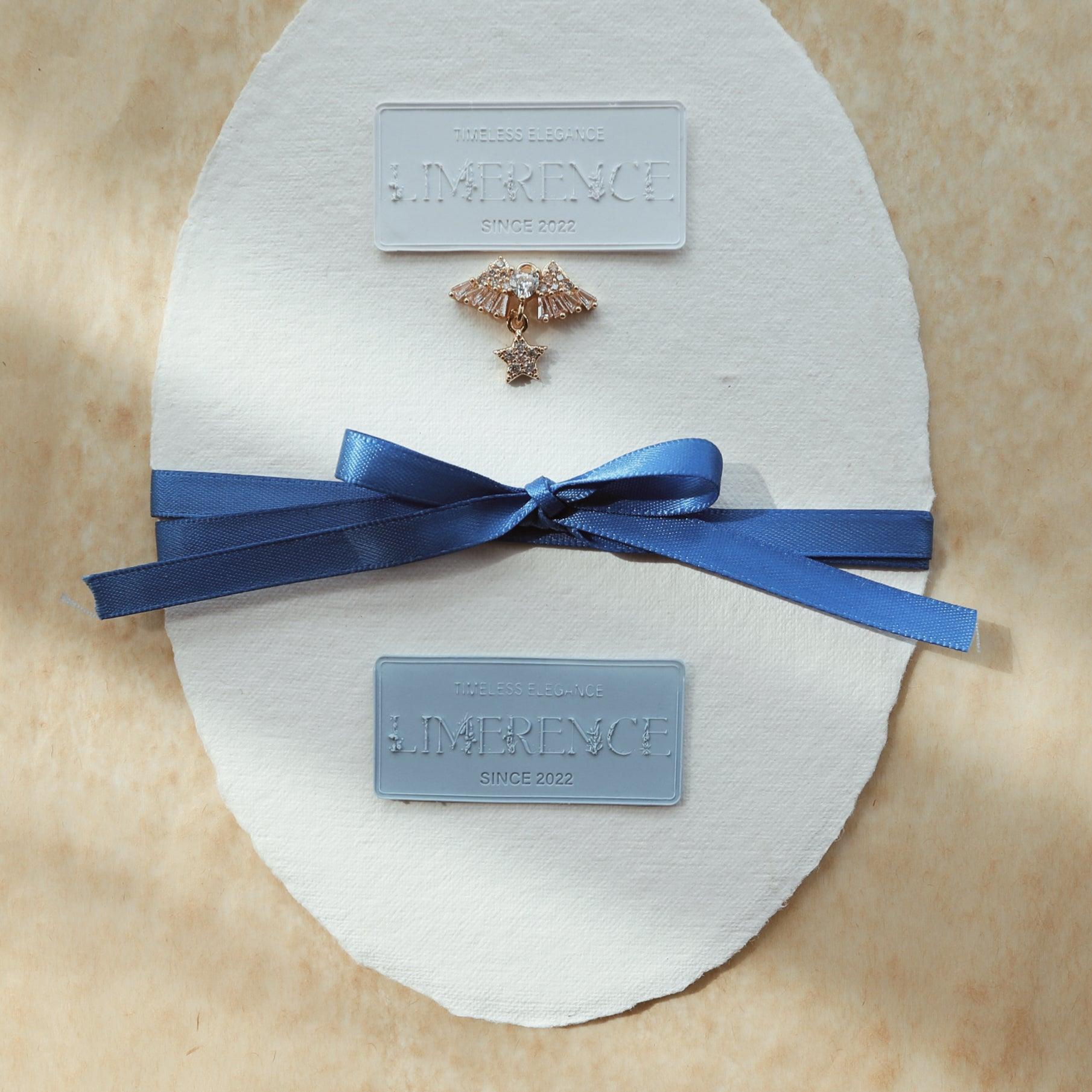 A sophisticated presentation featuring a light blue and a denim blue Limerence Seals label embossed with "TIMELESS ELEGANCE - LIMERENCE - SINCE 2022" on textured paper. A decorative gold pin and a blue satin ribbon complete the setup, highlighting the elegance and luxury associated with Limerence Seals, perfect for stylish invitations and high-end gift wrapping.