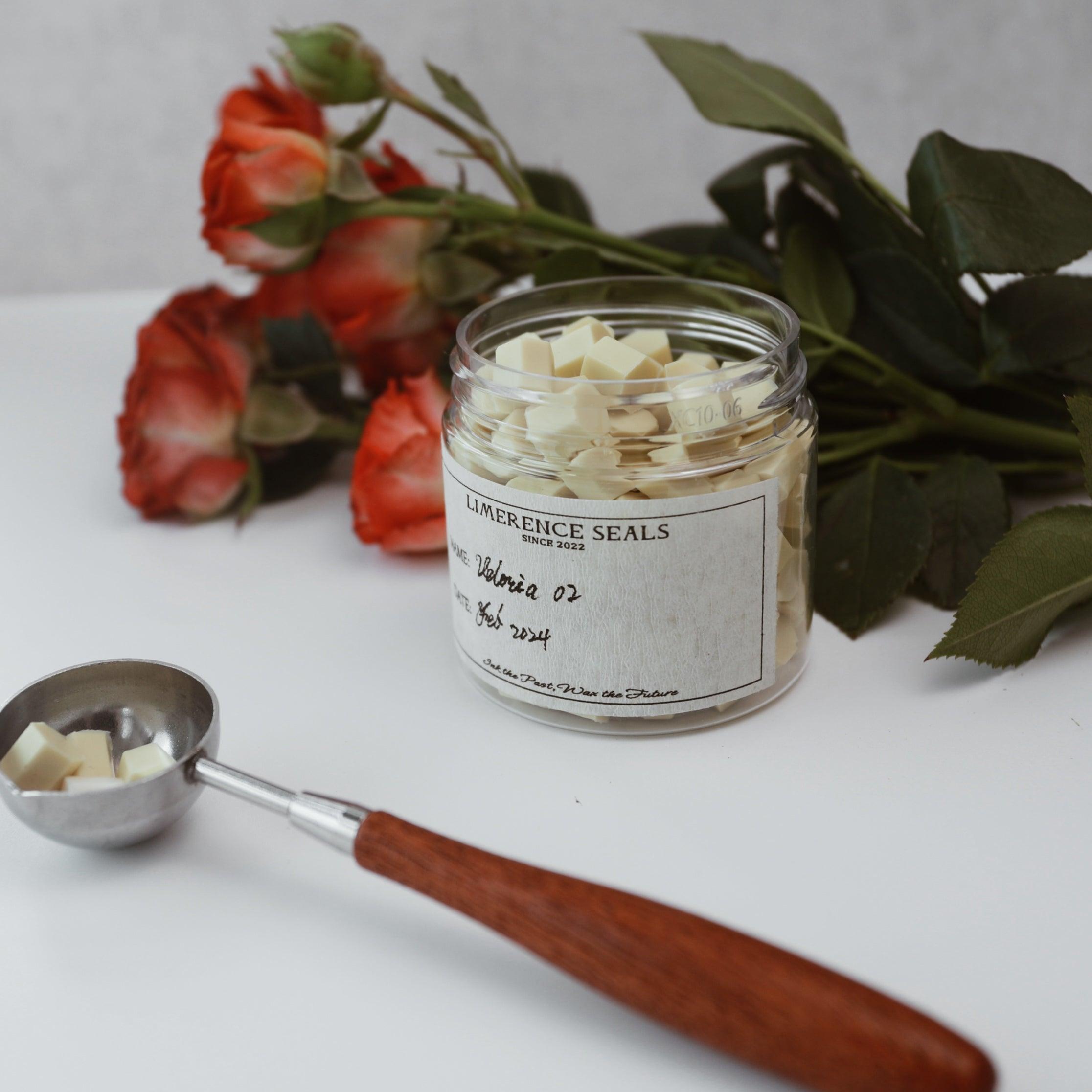 A jar of Veloria handmade sealing wax beads in ginger yellow by Limerence Seals, set against a backdrop of vibrant red roses. A wax spoon with a wooden handle holds a few beads, emphasizing the ease of use for crafting and sealing projects. This setup highlights the product's elegant design and suitability for adding a personalized touch to letters and gifts.